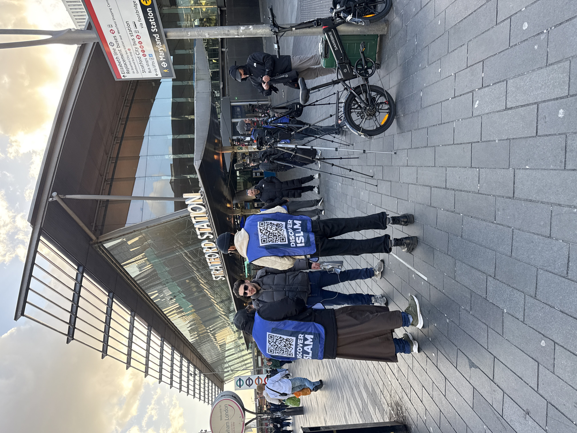 The image shows a scene outside Stratford Station in London, United Kingdom. The station entrance is visible, with a sign above it. A signpost indicates directions to Maryland Station and bus stops. Several people wearing blue vests with "DISCOVER ISLAM" printed on the back, along with a QR code, are standing on the pavement. A man stands near a bicycle, and several camera tripods are set up. The ground is paved with rectangular grey stones. The sky is visible in the upper left corner.