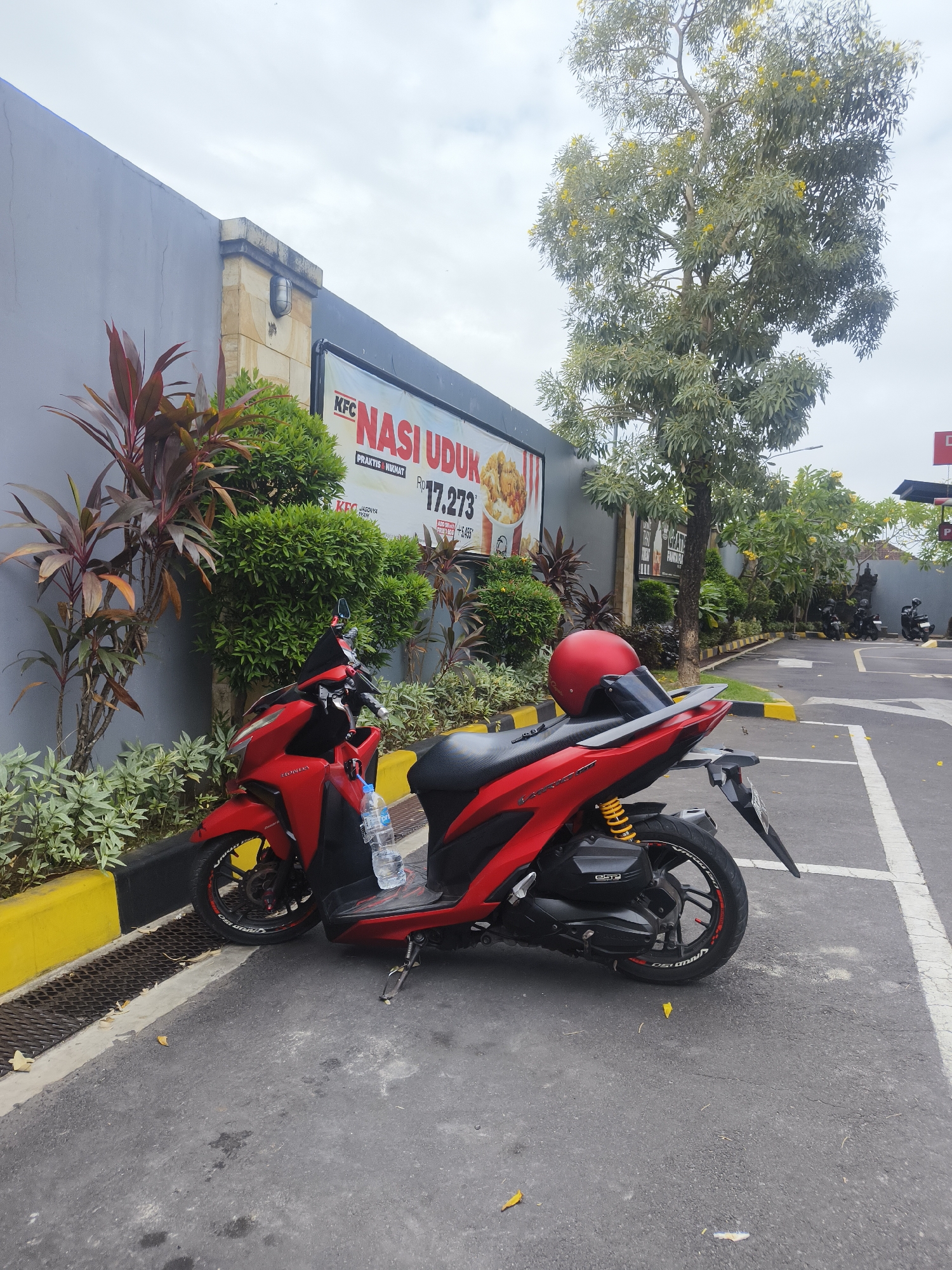 A red scooter is parked in front of a KFC restaurant in Denpasar, Indonesia. The scooter has a red helmet on its seat and a bottle of water in front of it. The KFC restaurant has a banner advertising "Nasi Uduk" for Rp 17,273. There are trees and plants around the restaurant, and other scooters are parked in the background. The sky is overcast.