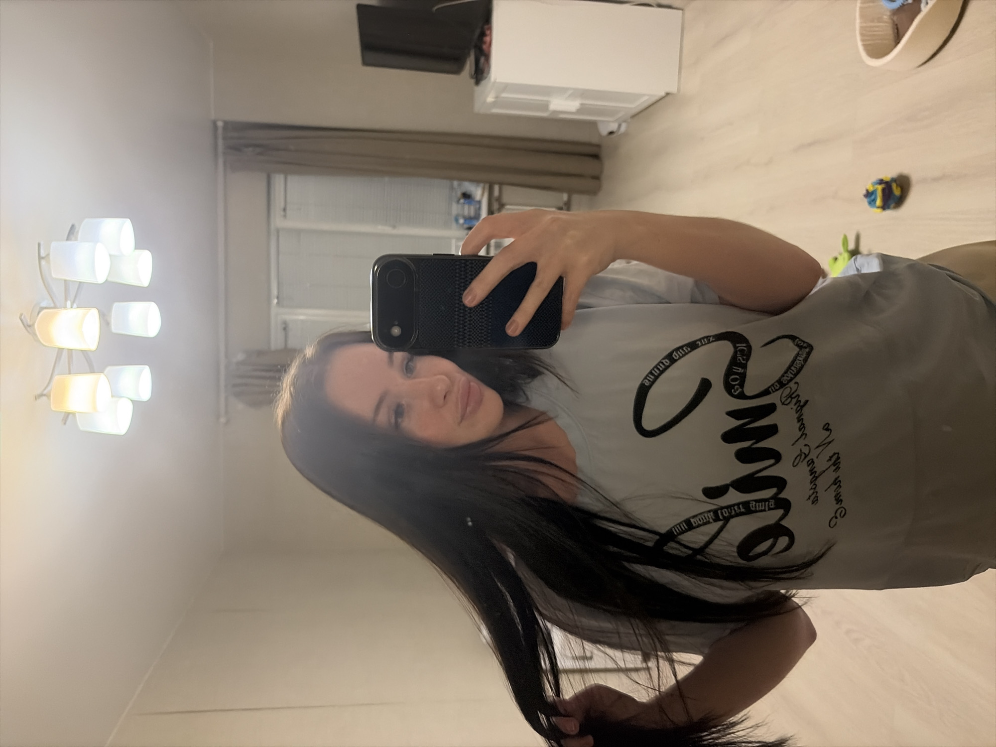 The image, rotated 90 degrees counter-clockwise, shows a young woman taking a mirror selfie indoors. She has long, dark hair and a neutral expression with a slight smile as she looks towards the camera reflected in the mirror. She is holding a dark-colored smartphone with a textured case in her right hand.
The woman is wearing a light gray t-shirt with black text on it. The most prominent word, when viewed upright, appears to be "Summer," written in a stylized script font. Smaller, less distinct text is also visible around this main word.
The background, visible through the mirror's reflection, reveals a domestic setting. On the left side, a modern floor lamp with multiple white and amber-glowing cylindrical shades illuminates the room. In the upper background, there's a white cabinet or dresser, and above it, a dark-colored television appears to be mounted on the wall. A window with horizontal blinds is also visible. The floor is covered with a light-colored wood laminate. On the floor in the reflection, near the bottom right, a small blue and yellow crinkled object, possibly a toy, and a green plush object are visible, alongside a light-colored wooden object that might be a pet bed or decorative tray. The overall lighting suggests artificial indoor lighting, possibly during the evening or night.