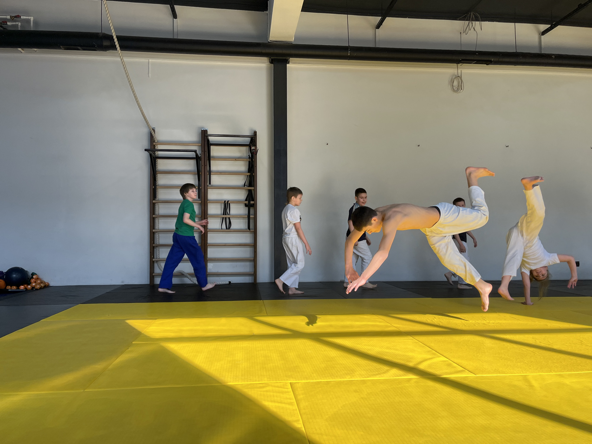 A group of children are practicing capoeira in a gym with yellow mats. One child is in the middle of a flip, another is performing a handstand, and others are standing or walking around. The gym has wall bars, a climbing rope, and a black ceiling structure. There are exercise balls on the side. The location may be Krasnoyarsk, Russia, but this cannot be verified from the image.