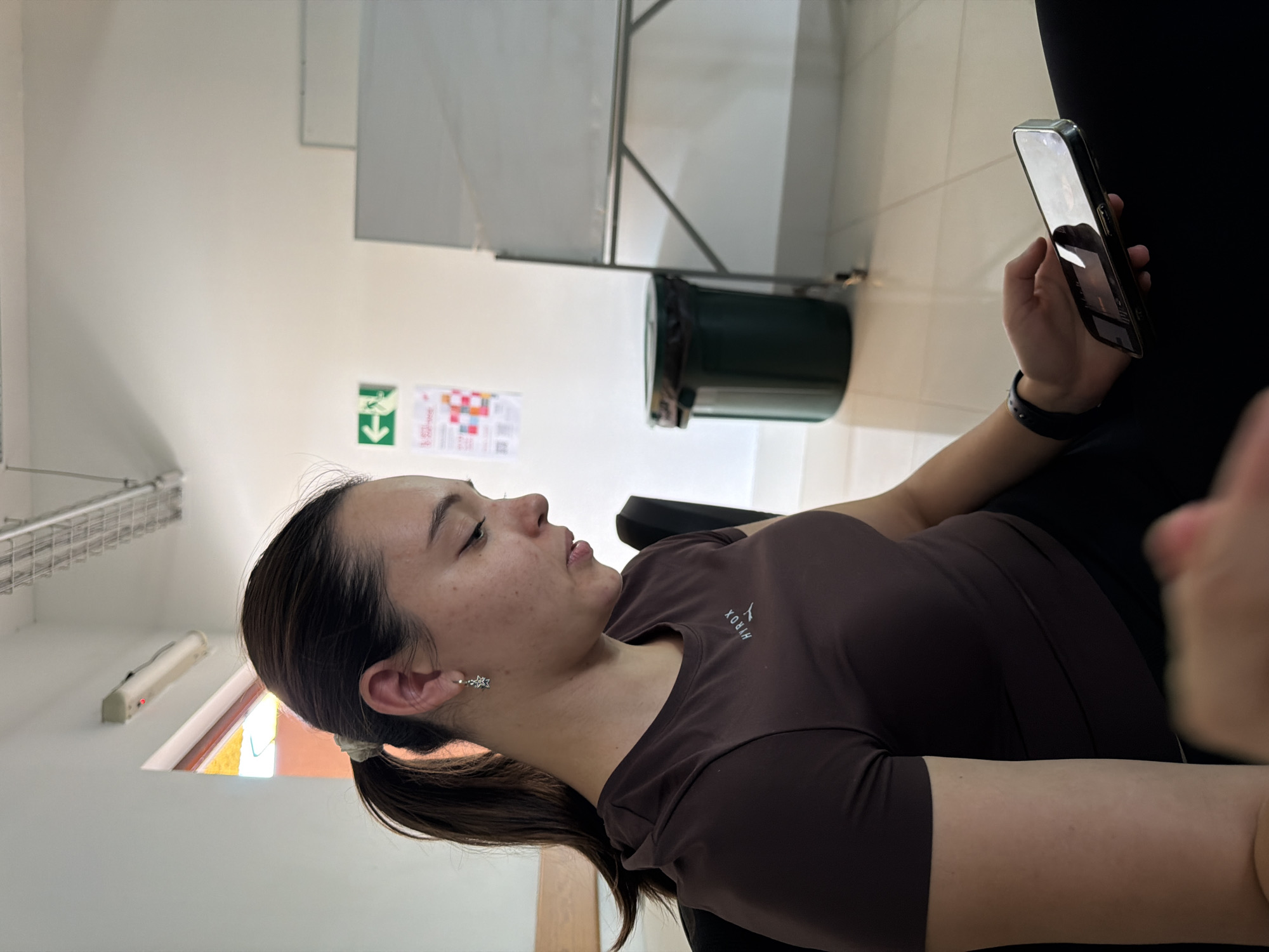 A young woman is seated, viewing a mobile phone. She is wearing a brown short-sleeved shirt with the word "HYROX" visible on the chest. Her hair is pulled back in a ponytail. The background reveals a wall with a green and white emergency exit sign, a sign with colored squares, a metal rack, and a green trash can. The location is San Andrés Cholula, Mexico.