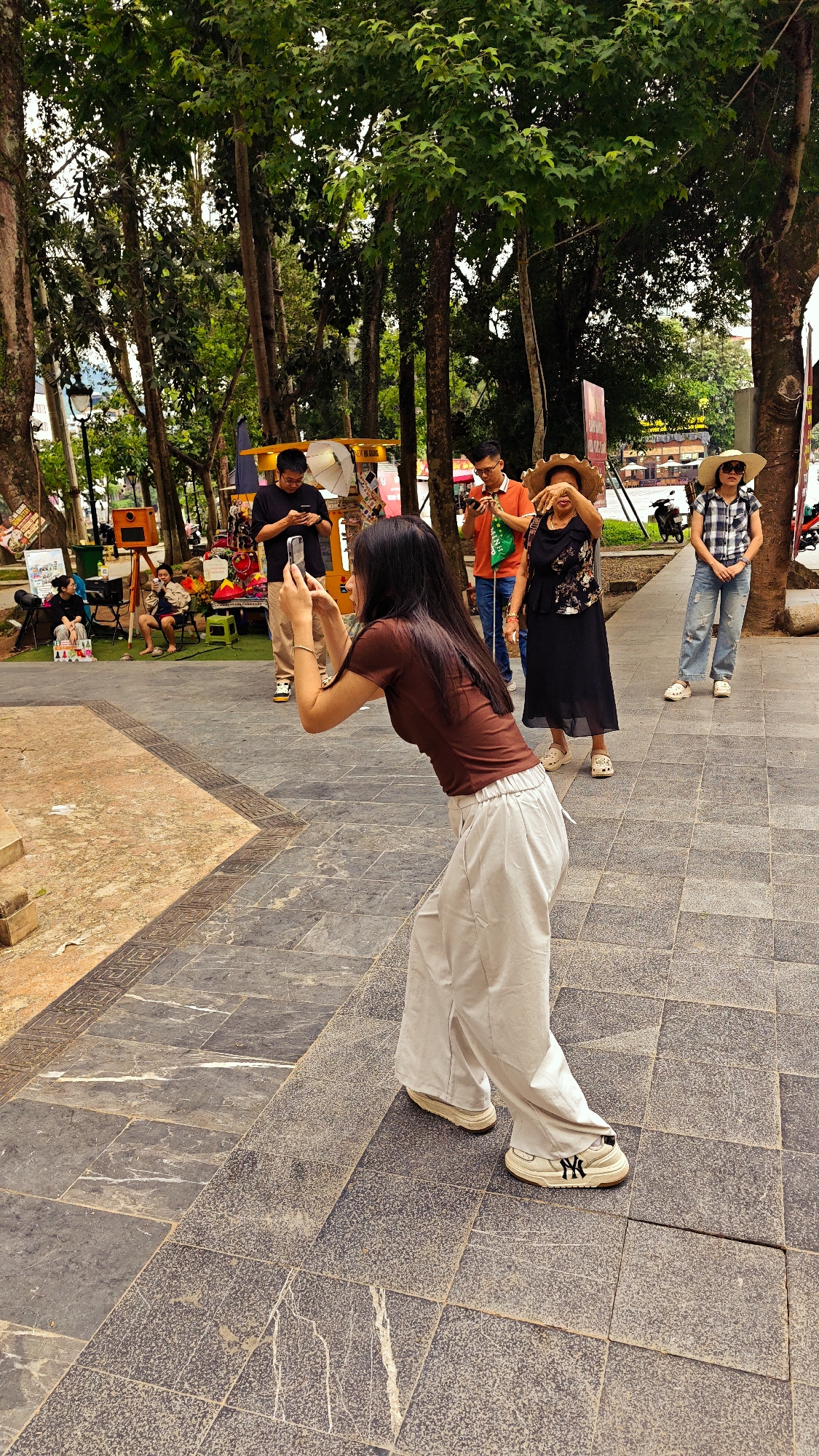 In a public park in the Hà Giang 1 Ward of Vietnam, a woman with long dark hair, wearing a brown top and light-colored pants, is in the foreground taking a picture with her smartphone. Behind her, three individuals stand on a tiled pathway. One man is using a phone, another man in an orange shirt appears to be taking a picture, and a woman in a black dress and straw hat is obscuring her face with her hand. A fourth person, wearing a plaid shirt and a straw hat, stands further back.
In the background, to the left, are several people sitting and relaxing, as well as a vendor stall. Trees surround the area, providing shade. The location appears to be an outdoor public space with pedestrian access.