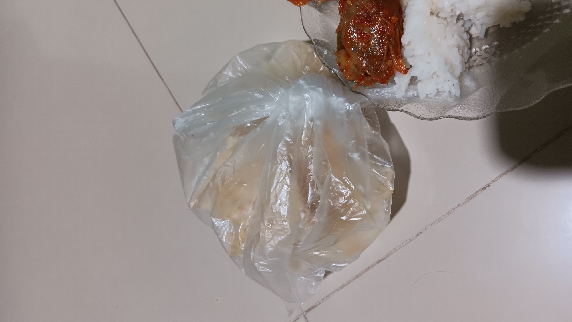A plastic bag containing pale, flat food items is situated on a tiled floor. To the right of the bag, a clear, wavy-edged bowl holds white rice and a piece of meat coated in a red sauce. The lighting suggests an indoor setting. The location is identified as Sonobekel, Indonesia.