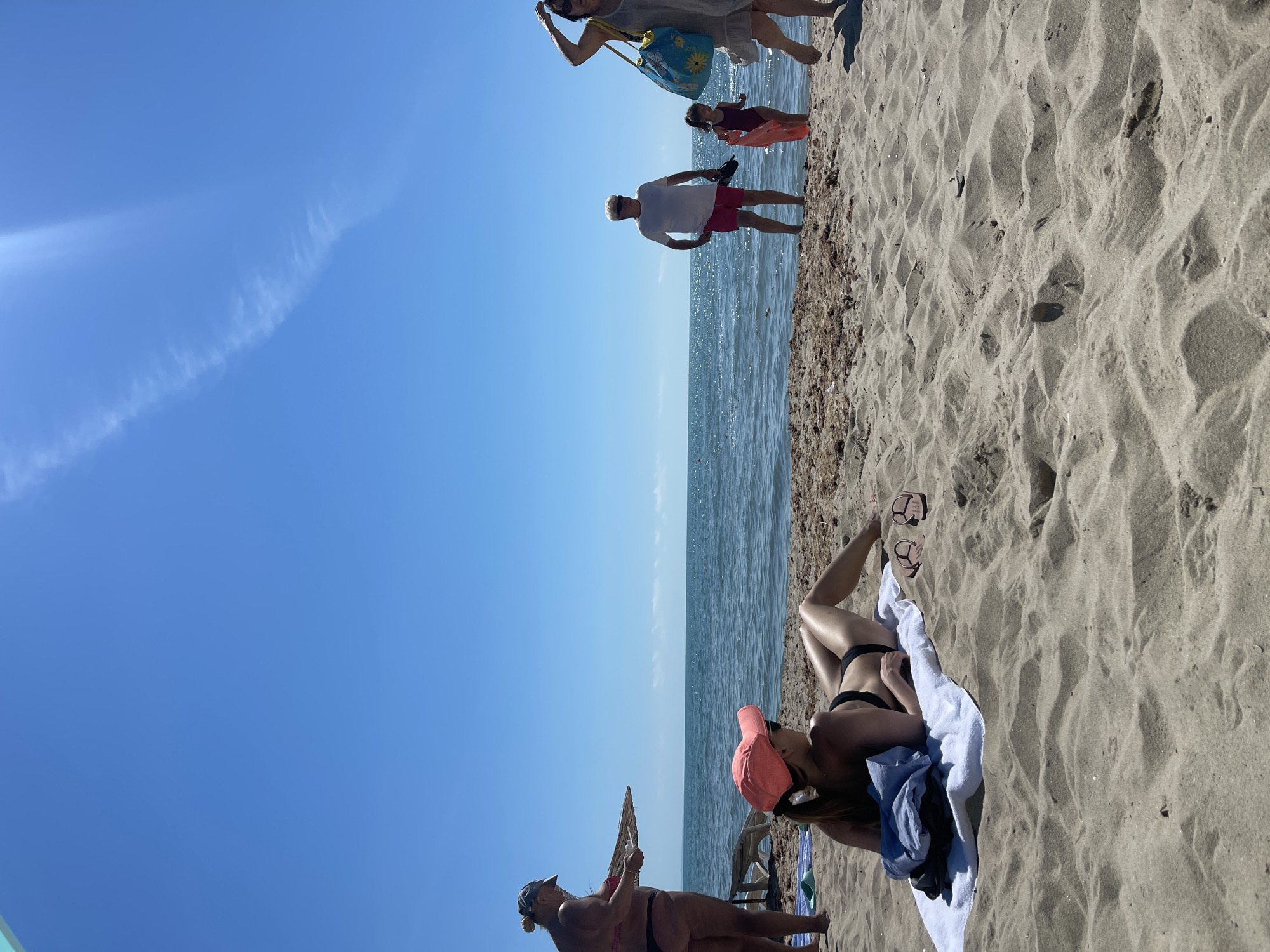 The image depicts a sunny day at the beach in Kranevo, Bulgaria. Several people are visible enjoying the seaside. In the foreground, a person in a black bikini is lying on a white towel, with a light blue shirt and dark clothing placed nearby. Another person, wearing a pink cap and a bikini, is seated next to them, holding a straw sunshade. Further up the beach, a man in a white t-shirt and red shorts stands near the water's edge, holding a black bag. A young child in a red swimsuit is also present, holding an orange inflatable toy. At the top of the frame, a person with sunglasses and a light-colored dress is seen carrying a colorful tote bag. The sea is calm and stretches to the horizon, under a clear blue sky with a few wispy clouds. The sand is a light beige, marked with the distinct patterns of ripples. The overall scene suggests a relaxed and leisurely day at the beach.