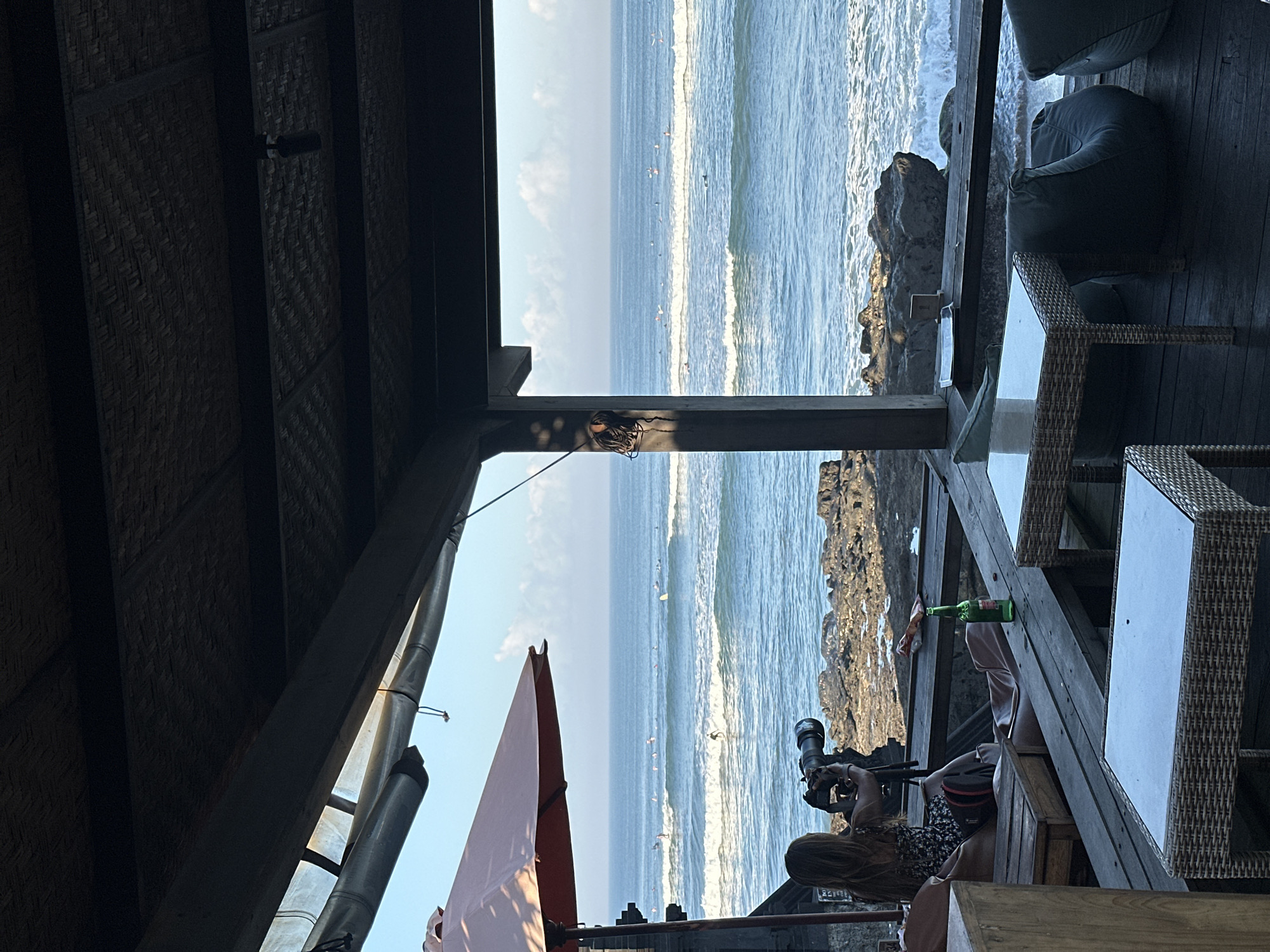 The image depicts a scene from a beachfront establishment in North Kuta, Indonesia, likely a restaurant or bar with an outdoor seating area. The perspective is from beneath a thatched roof structure, looking out towards the ocean.
In the foreground, a woman with long, brown hair is seated at a table, holding a professional camera with a large lens. She appears to be focused on taking photos or videos of the ocean. Beside her, a green bottle of Bintang beer rests on the wooden table. The seating area features outdoor furniture, including wicker chairs and tables, as well as large, cushioned beanbag chairs visible in the background.
The ocean stretches out to the horizon, with moderate waves breaking on a rocky shore. Numerous seabirds are scattered across the water and the sky, suggesting a natural, coastal environment. The sky is a clear blue with some scattered white clouds.
The lighting suggests it is daytime, likely late afternoon or early evening, given the warm tones and shadows. The overall atmosphere seems relaxed and scenic, typical of a tropical beach destination. The presence of the camera indicates a potential interest in wildlife, surf, or the general beauty of the location. The orientation of the image is sideways, with the horizon running vertically.