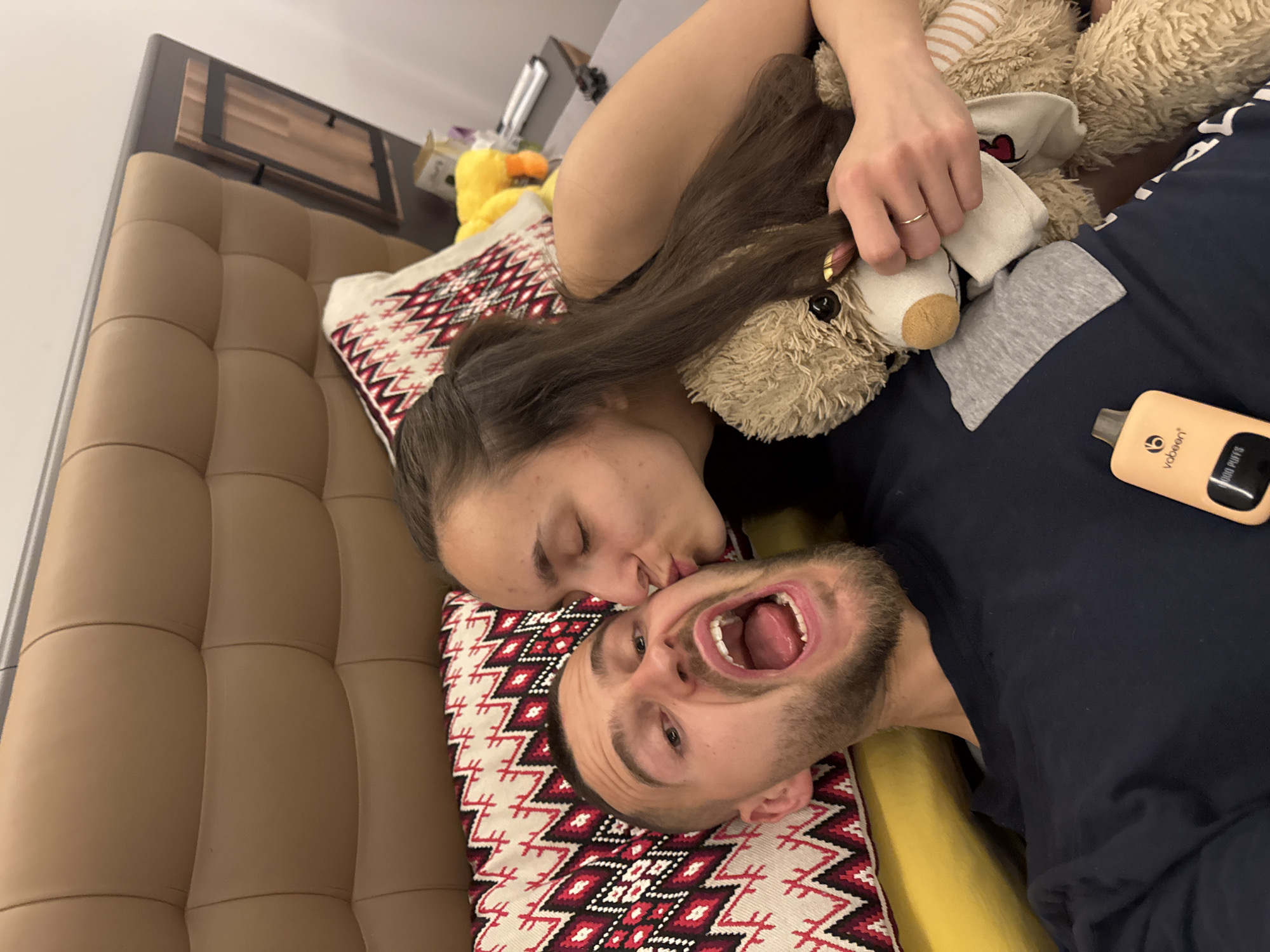 A woman is kissing a man on the cheek. The woman has long, brown hair. The man has short, brown hair and a beard. He is smiling with his mouth open, showing his teeth. They are lying on a bed with a decorative pillow. The woman is holding a teddy bear. A yellow rubber duck is near the teddy bear. A vape pen is visible near the man. The setting appears to be indoors. There are no identifying features to locate the scene within Sofia, Bulgaria.