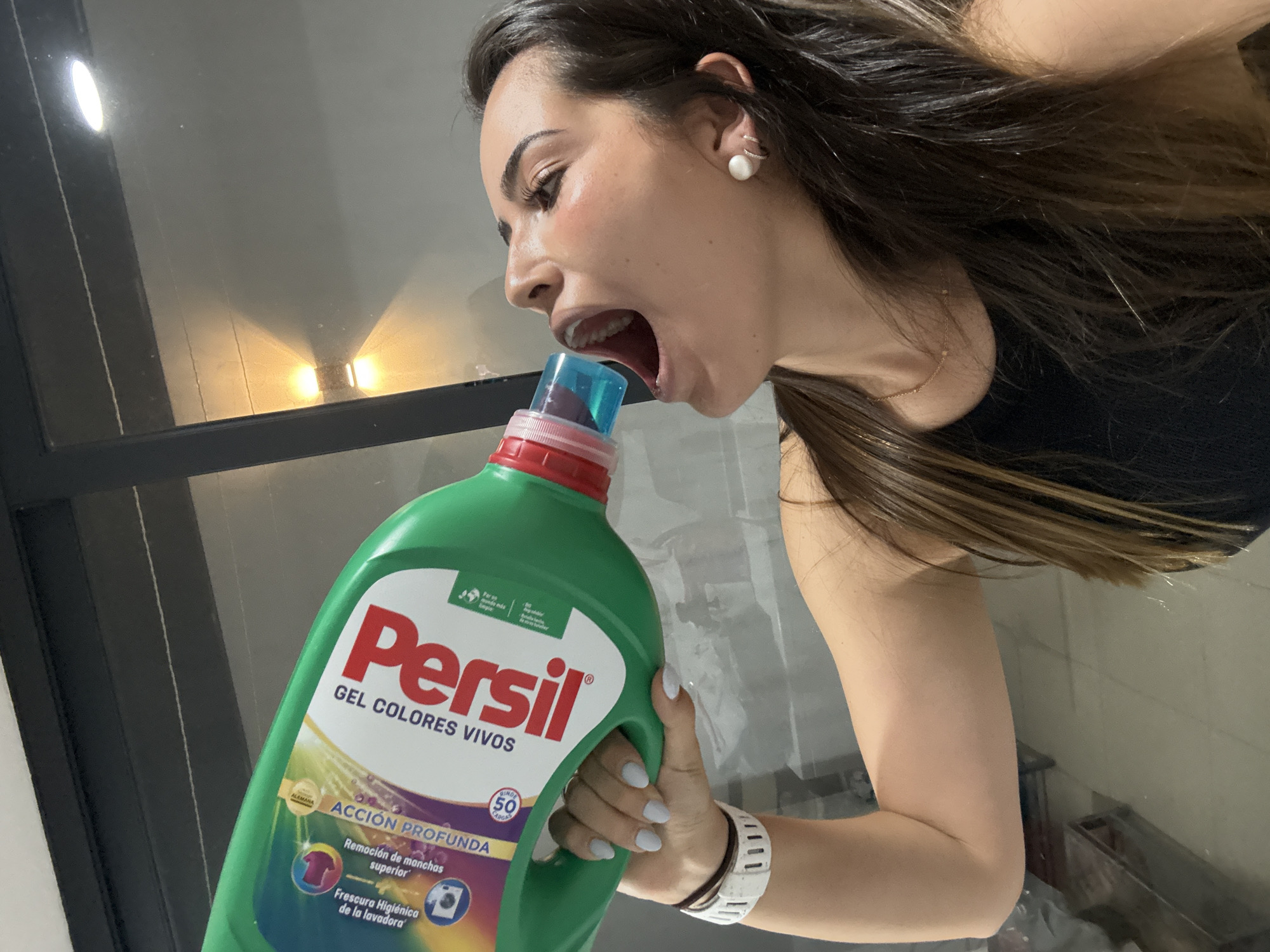 A woman is holding a green bottle of Persil Gel Colores Vivos laundry detergent and placing the blue nozzle in her mouth. The brand is visible. She is wearing a white wristwatch and pearl earrings. She has long brown hair. The background shows a window frame and a tile wall. The location is unspecified but might be a home or apartment. The city of San Bernardino Tlaxcalancingo, Mexico, is not visible.