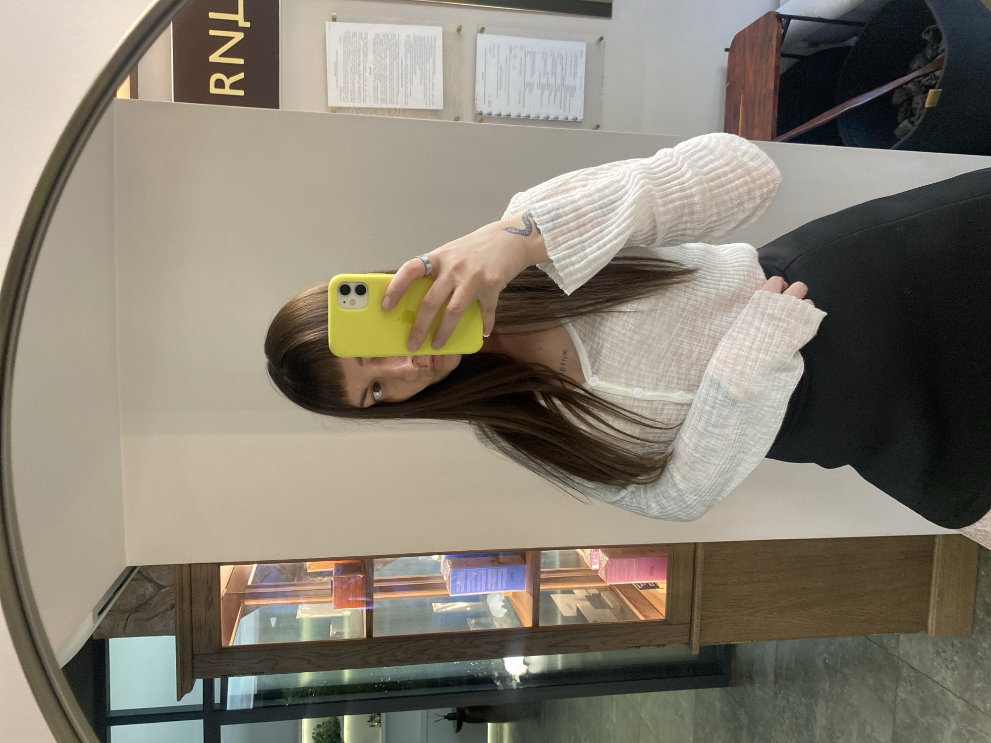 A woman is taking a selfie in a mirror. She is holding a yellow phone. She is wearing a white shirt and a black skirt. The background includes a cabinet with glass doors displaying various items, signage with the letters "RNL," and framed documents on the wall. The location is possibly an interior space in the city of Votkinsk, Russia.