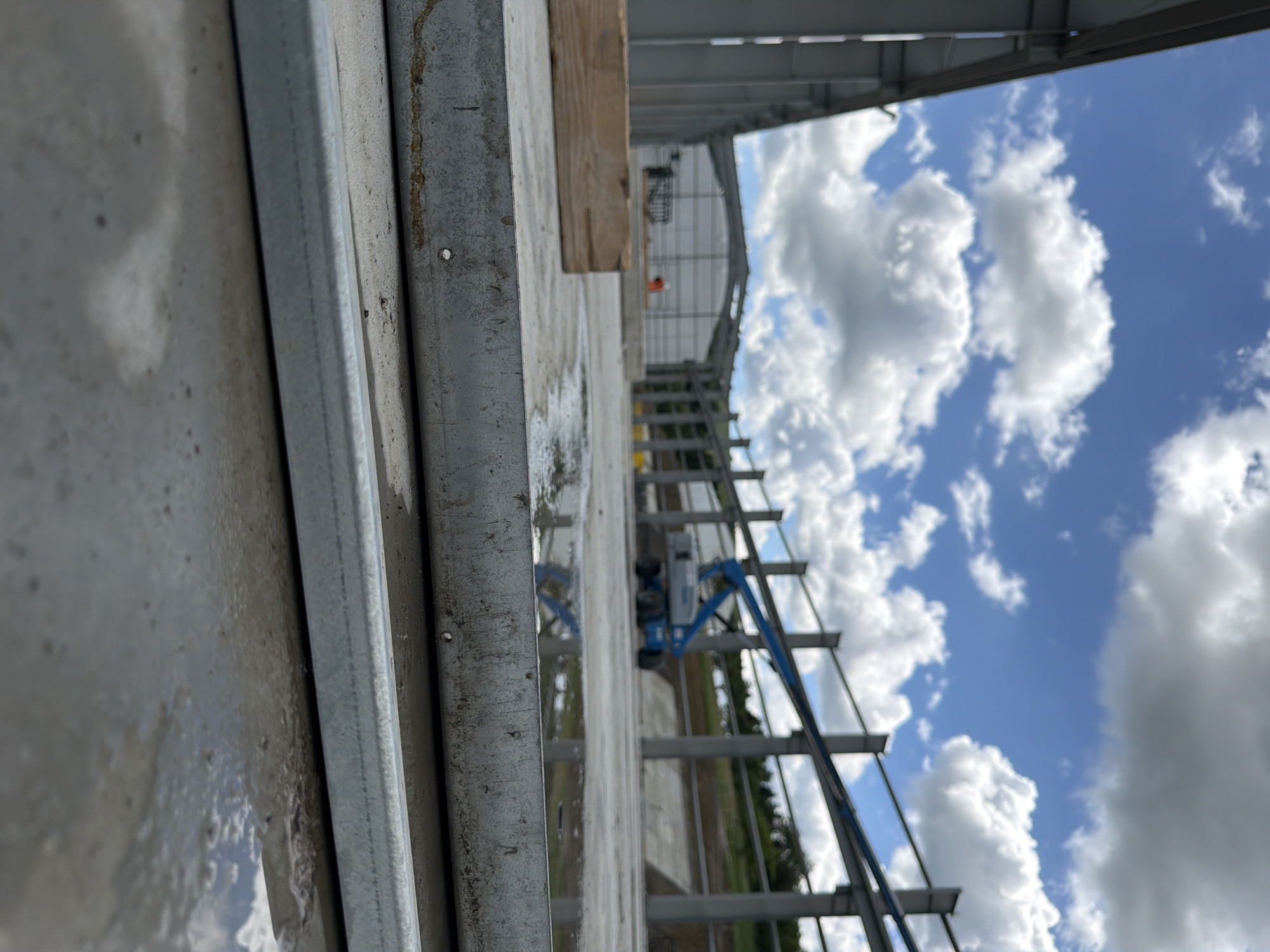 A construction site in Janowice, Poland, displays the initial stages of a building's frame. The foreground is dominated by a wet concrete surface, with two horizontal metal beams resting on it. The beams show signs of wear, with some rust and dirt accumulation. To the left, a rectangular piece of weathered wood lies on the concrete.
In the mid-ground, the skeletal structure of a building is taking shape, with vertical steel columns supporting horizontal beams. A blue boom lift, a piece of heavy machinery used for elevated access, is parked near the structure. The area surrounding the construction is partially covered with concrete, with some sections appearing wet and reflecting the sky.
The background features a large, partially completed building with a white, corrugated metal exterior. Further in the distance, a tree-lined slope rises, and the sky is filled with a mix of white, puffy clouds against a bright blue expanse. Two individuals, one wearing an orange vest, are visible near the partially completed building in the distance, suggesting ongoing construction activity. The overall scene depicts an active construction zone under a bright, cloudy sky.