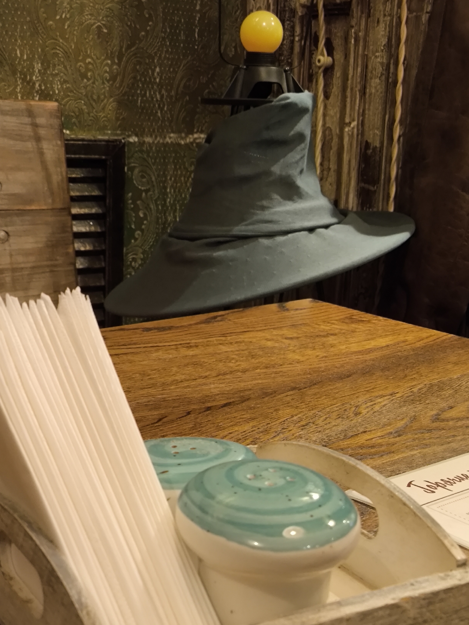 A close-up shot depicts a wooden table with a Sorting Hat replica leaning against a wall decorated with patterned wallpaper. A tray with napkins and salt and pepper shakers sits on the table in front of the hat. A lamp with a yellow bulb is positioned near the hat. The location is Moscow, Russia.