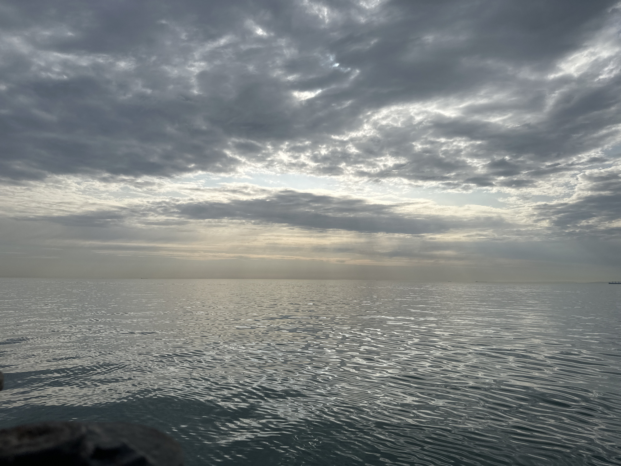 The image depicts a body of water, likely a sea or large lake, under a sky filled with dense, gray clouds. Light penetrates through breaks in the cloud cover, creating a diffuse glow on the horizon and reflecting on the water's surface. The water is relatively calm, with small waves visible. The horizon is distant, with faint outlines of ships or structures visible. The location is identified as Unknown, Türkiye. No people are visible in the image. A dark object is in the lower left corner of the image that appears to be a part of the boat from where the photo was taken.