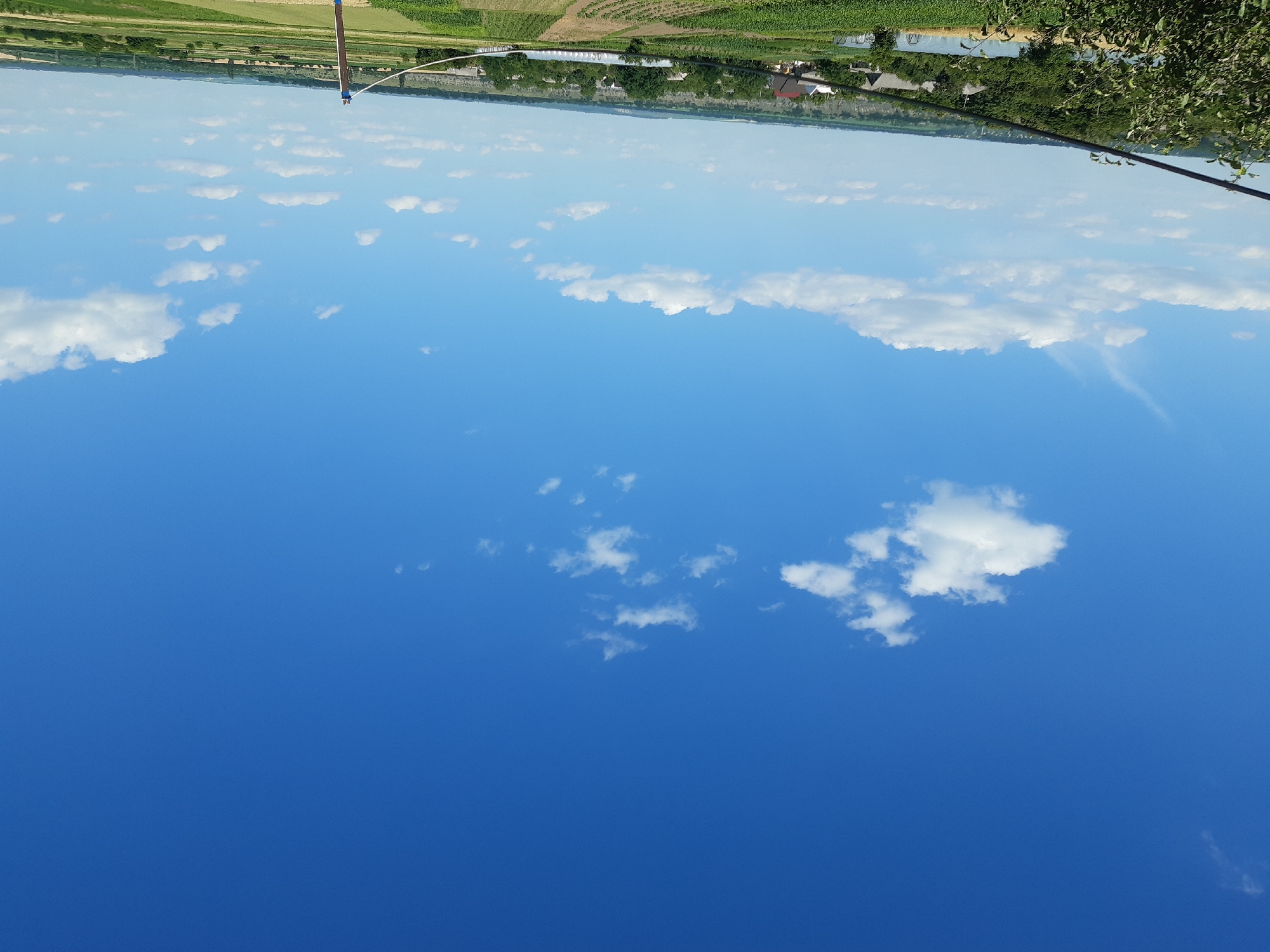 The image shows a wide, clear blue sky with scattered white clouds. In the distance, a green landscape is visible, featuring fields, trees, and some buildings. The scene is oriented sideways, suggesting the photo was taken in portrait mode and then rotated. The overall impression is of a bright, sunny day, likely during the daytime, given the strong blue of the sky and the visibility of the landscape. The location is identified as Kavetchyna, Ukraine. No people or specific activities are discernible, but the scene evokes a sense of open space and nature.