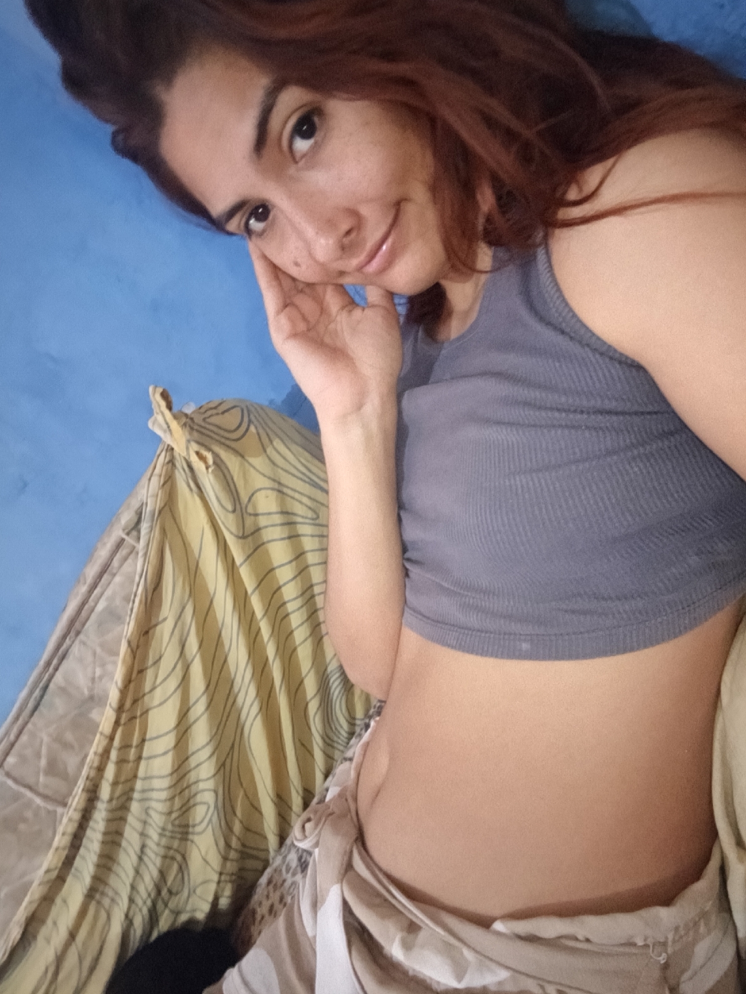 A woman with auburn hair and fair skin is lying in a hammock, wearing a gray cropped tank top and patterned pants. She is propped up, resting her head on her left hand and smiling. The hammock is beige with yellow patterned fabric and hangs against a blue wall. The location is unknown.