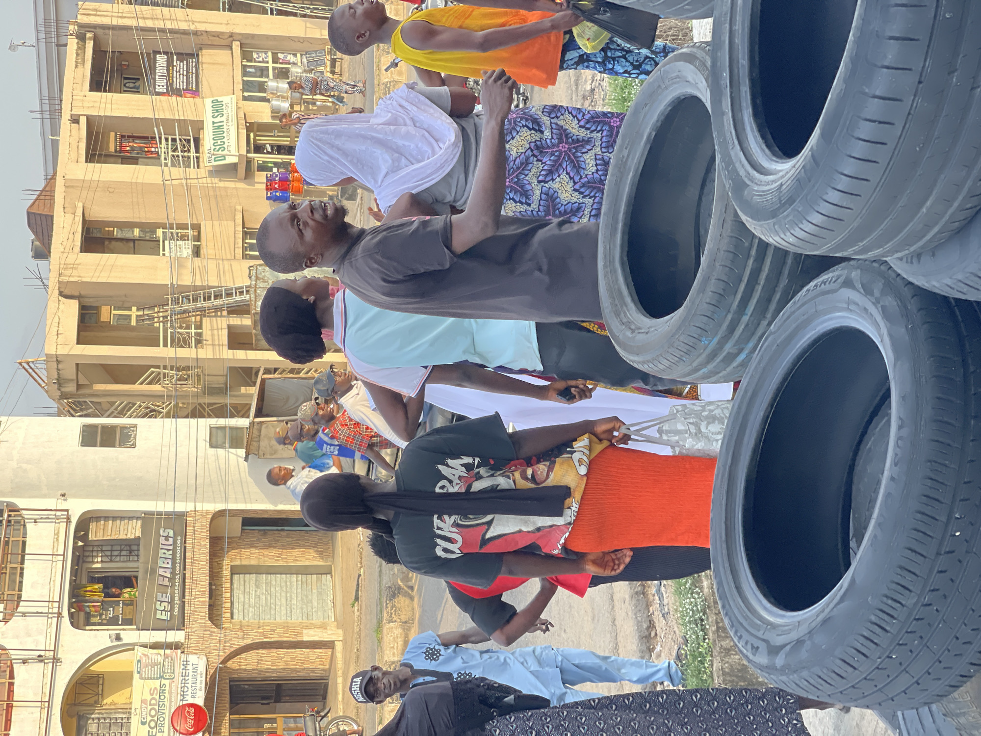 In an outdoor urban environment, a group of people are gathered near a street. In the foreground, several large tires are stacked. To the right, a young Black male wearing a yellow and orange tank top and blue patterned pants stands, holding a black plastic bag. To his left, a Black female wearing a white hijab and a patterned skirt stands facing away from the camera. To her left, a Black male in a dark t-shirt and a Black female with a black headscarf and an orange skirt are facing away from the camera. In the background, several buildings are visible, including one with signs for "ESE FABRICS" and "MOREMI RESTAURANT". Another building has a sign for "BEAUTYRYMD" and "D'SCOUNT SHOP". People are visible inside the buildings and walking on the street. A motorcycle is parked on the left side of the image. The lighting suggests it is daytime.