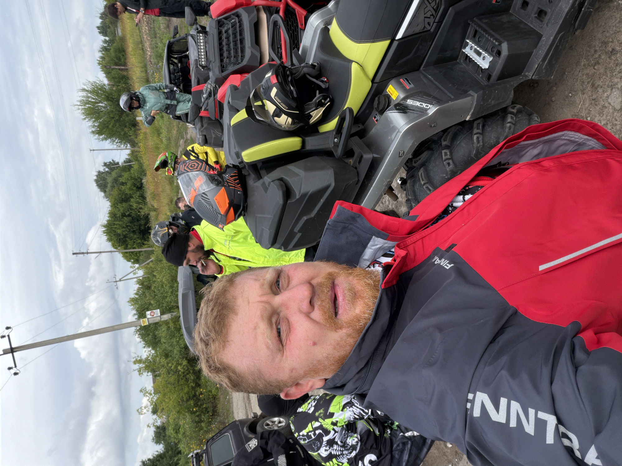 In Kharkiv, Ukraine, a man with red hair and a beard, wearing a red and grey jacket, is the primary subject of the image, positioned in the foreground and looking directly at the camera. Behind him, several ATVs are parked, some with helmets placed on their seats. One ATV has the brand name "CAN-AM" visible. To the left of the man, another person in a black and green patterned jacket is partially visible, and a black car is also present. In the background, a line of trees and a cloudy sky are observed, with utility poles and wires suggesting an outdoor, possibly rural or semi-rural, setting on the outskirts of the city. The overall scene depicts a group preparing for or concluding an ATV activity.