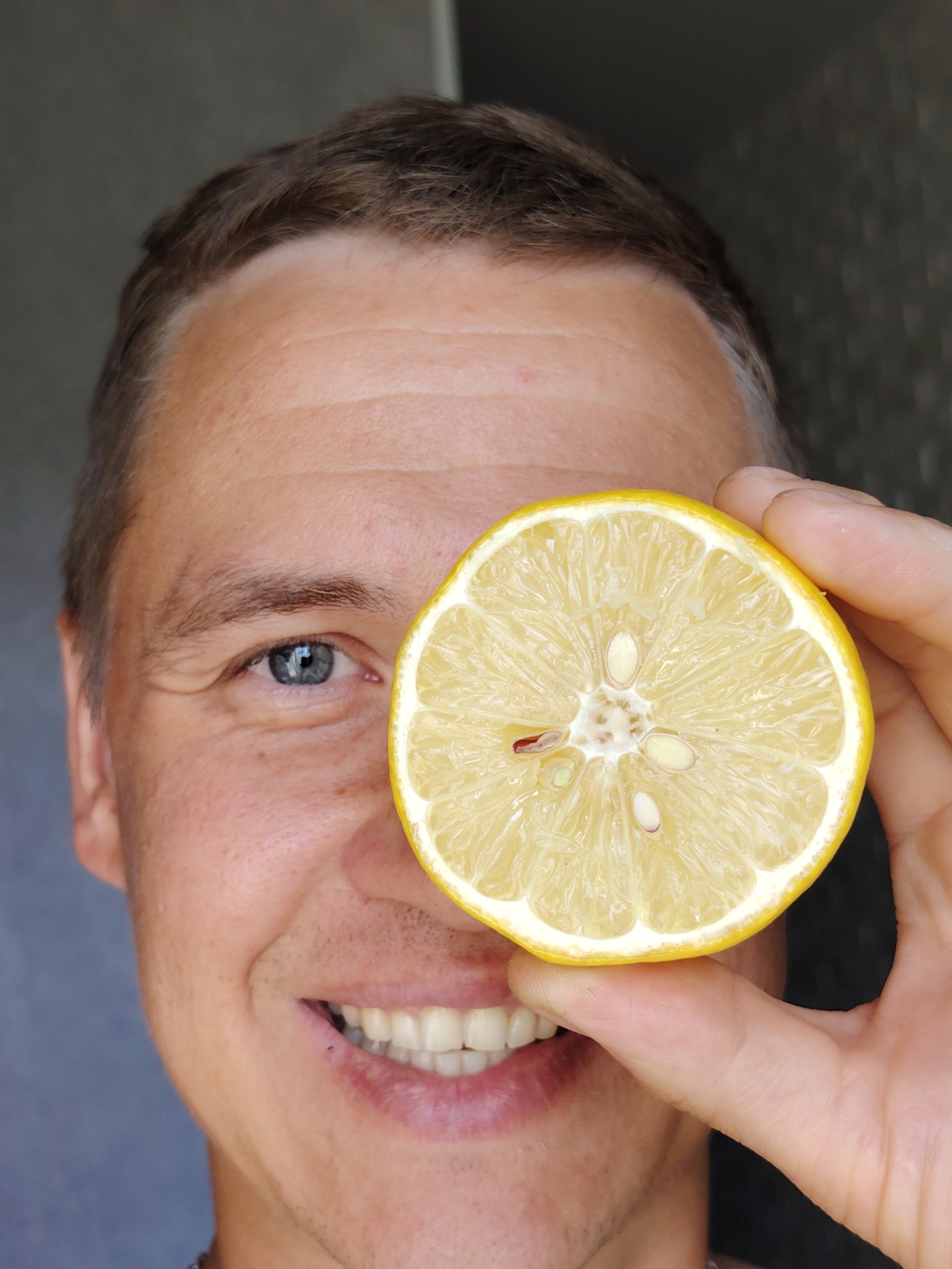 A man's face is shown close-up, with a slice of lemon covering his right eye. He is smiling broadly, revealing his teeth. His left eye is visible, and it is blue. The man has short, brown hair and light skin. The background is a blurred, neutral gray. The image appears to be taken outdoors, possibly in a residential area. There is no specific information about the location in the city of Chaikovsky, Russia, visible in the image.