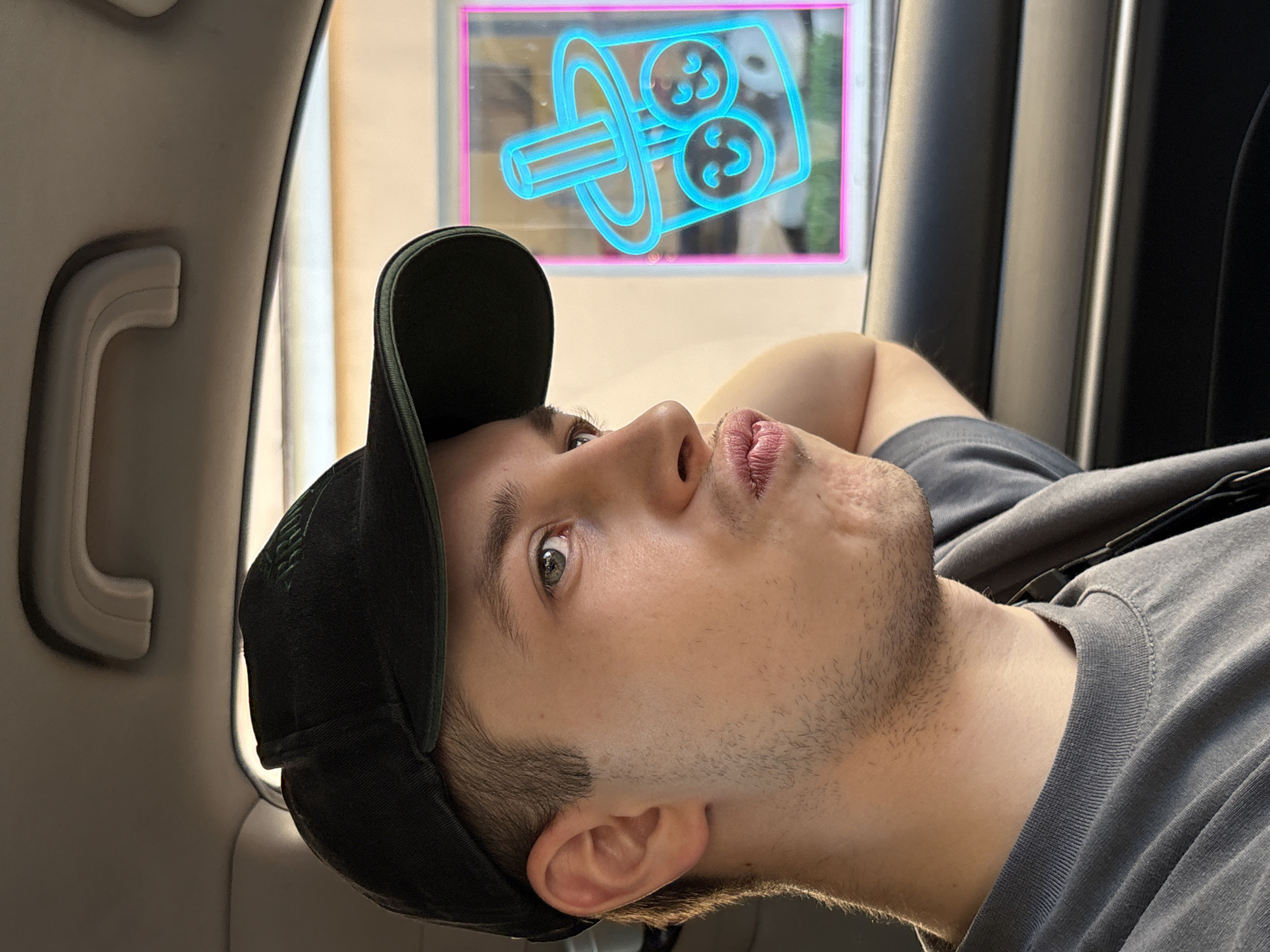 The image displays a young adult male in what appears to be the front passenger seat of a vehicle, looking towards the right side of the frame, out a window. He is wearing a dark-colored baseball cap with an illegible light-colored embroidered design on the front, and a dark grey crew-neck t-shirt. The subject has short light brown hair, visible stubble on his jawline and chin, and light-colored eyes. His lips are pursed in a distinct pout. The car's interior visible elements include a light-colored headliner with a white grab handle and a dark-colored door panel with a horizontal accent line. Outside the car window, a light-colored building is visible, featuring a prominent neon sign outlined in pink. The sign itself is light blue and depicts a bubble tea cup with a straw and two smiling, spherical elements within. The lighting suggests daytime. The exact location within Saint Petersburg, Russia, is not identifiable from the visual information presented.
