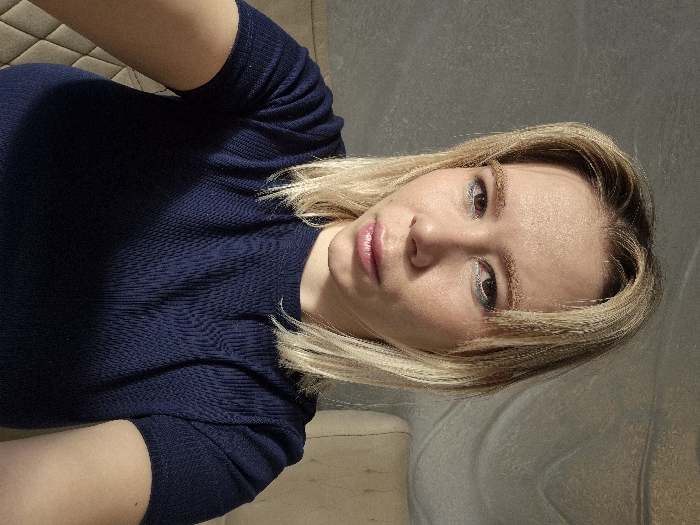 A light-skinned woman with blonde hair is wearing a dark blue textured knit top. She is looking directly at the camera with a slight pout. She appears to be in a room with light-colored walls and flooring. The image is rotated slightly, and the overall lighting is bright.