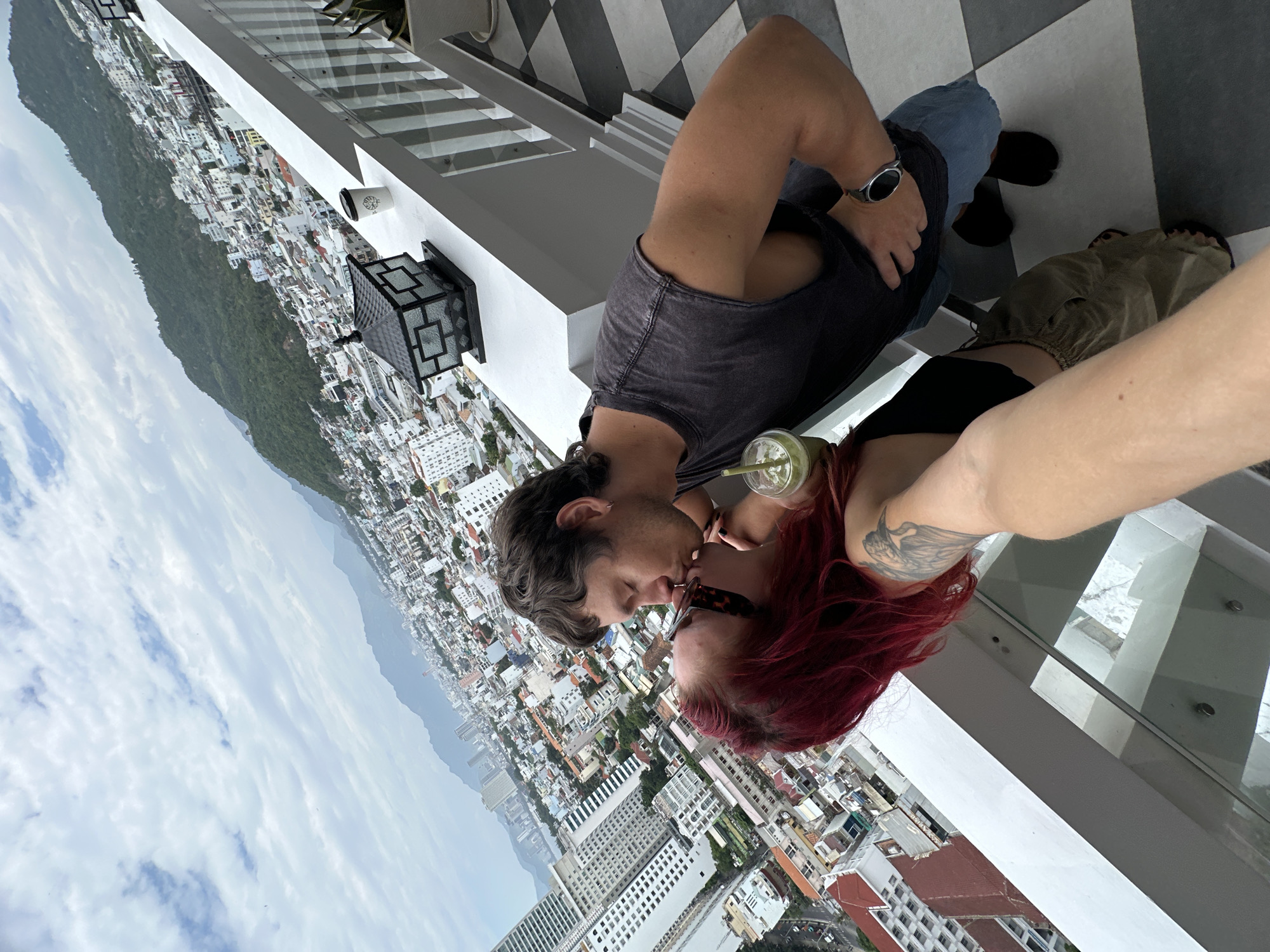 A couple kisses on a balcony in Nha Trang, Vietnam, with the city and mountains visible in the background. The man wears a dark tank top and denim shorts, while the woman has red hair and wears sunglasses and a black top. A green drink sits on the balcony railing. The balcony has a checkered tile floor and a white railing.
