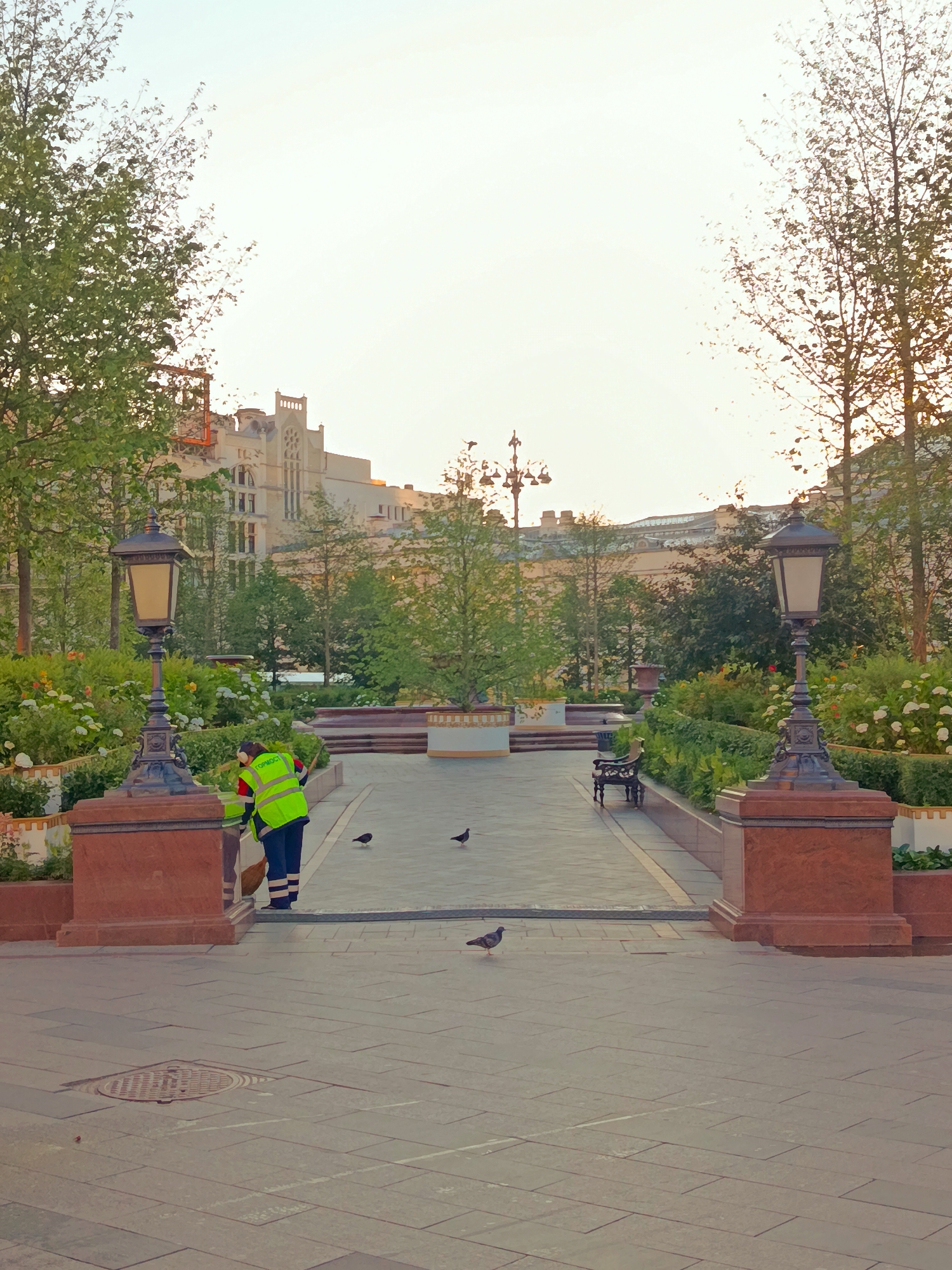 In an urban park setting, a sanitation worker wearing a high-visibility yellow vest and dark pants is sweeping the paved ground with a broom. The worker is positioned between two ornate lampposts and is facing away from the camera. Several pigeons are scattered across the pathway. In the background, there are trees, flowerbeds, a park bench, and a large, ornate building with a pale facade. The sky is clear and bright.