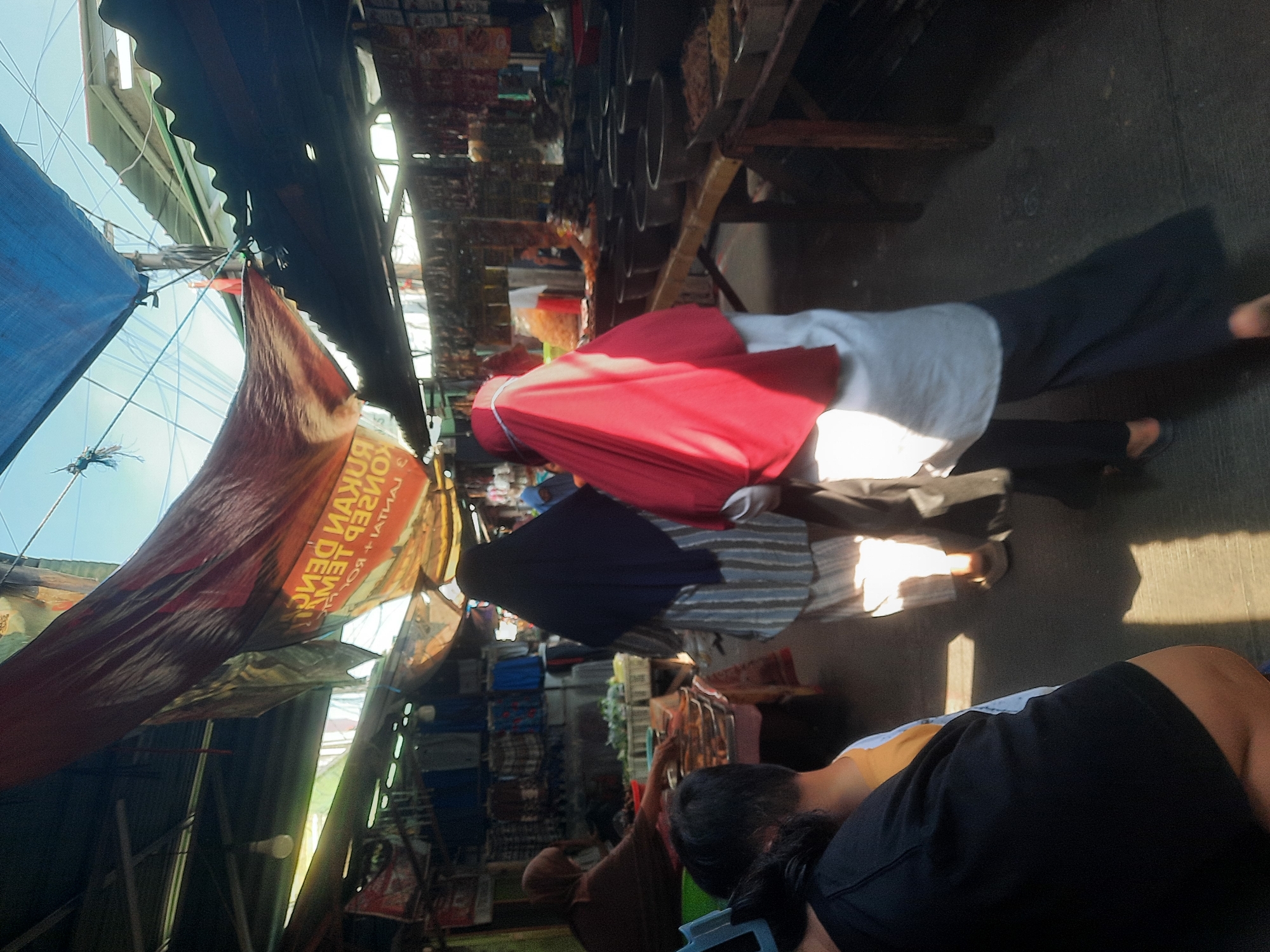 This image captures a bustling outdoor market scene in North Jakarta, Indonesia, likely a traditional wet market or a roadside vendor area. The foreground shows the backs of several people, some wearing headscarves. A person in the center is seen from behind, wearing a dark hijab and a striped outfit, while another person to their right is wearing a bright red hijab and a white top. The market is covered by a patchwork of tarps and corrugated metal roofing, allowing shafts of sunlight to illuminate the pathway. Stalls are set up on either side, displaying various goods. On the left, one stall appears to be selling fresh produce, with a vendor in a brown hijab reaching towards it. On the right, a long wooden table is laden with numerous round containers filled with what seems to be dry goods, spices, or other market products. The overall atmosphere suggests a busy commercial area with a variety of goods on display and people engaged in shopping or selling.