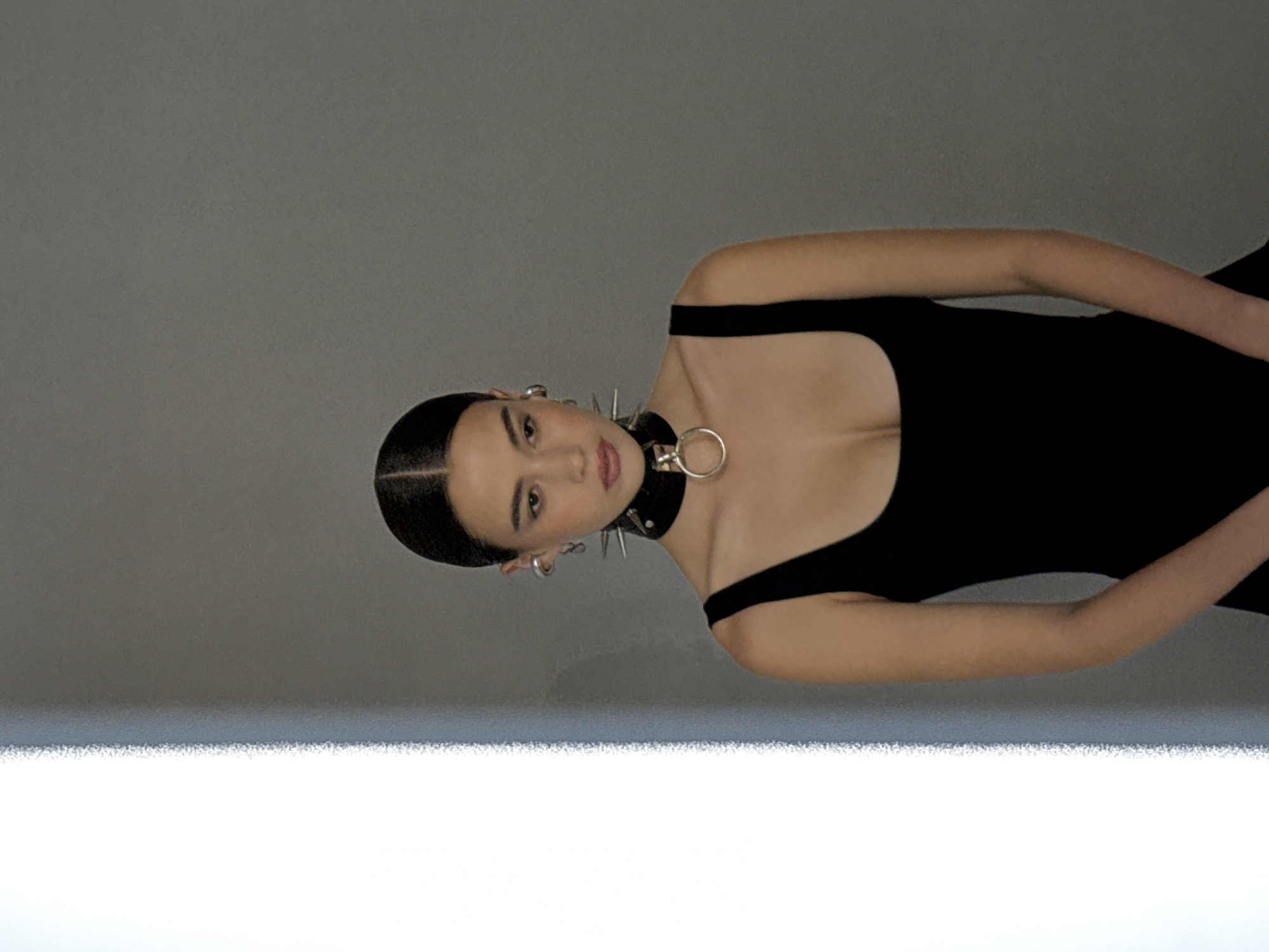 The image shows a young woman wearing a black sleeveless leotard and a spiked choker necklace. She has dark hair pulled back neatly, and she is wearing large hoop earrings. She is standing in front of a grey background with a bright white vertical light source on the left side of the image. The lighting suggests a studio setting, possibly in an area of London, United Kingdom. There are no visible indicators that place the scene specifically within the city of London beyond the possibility of a studio setting.