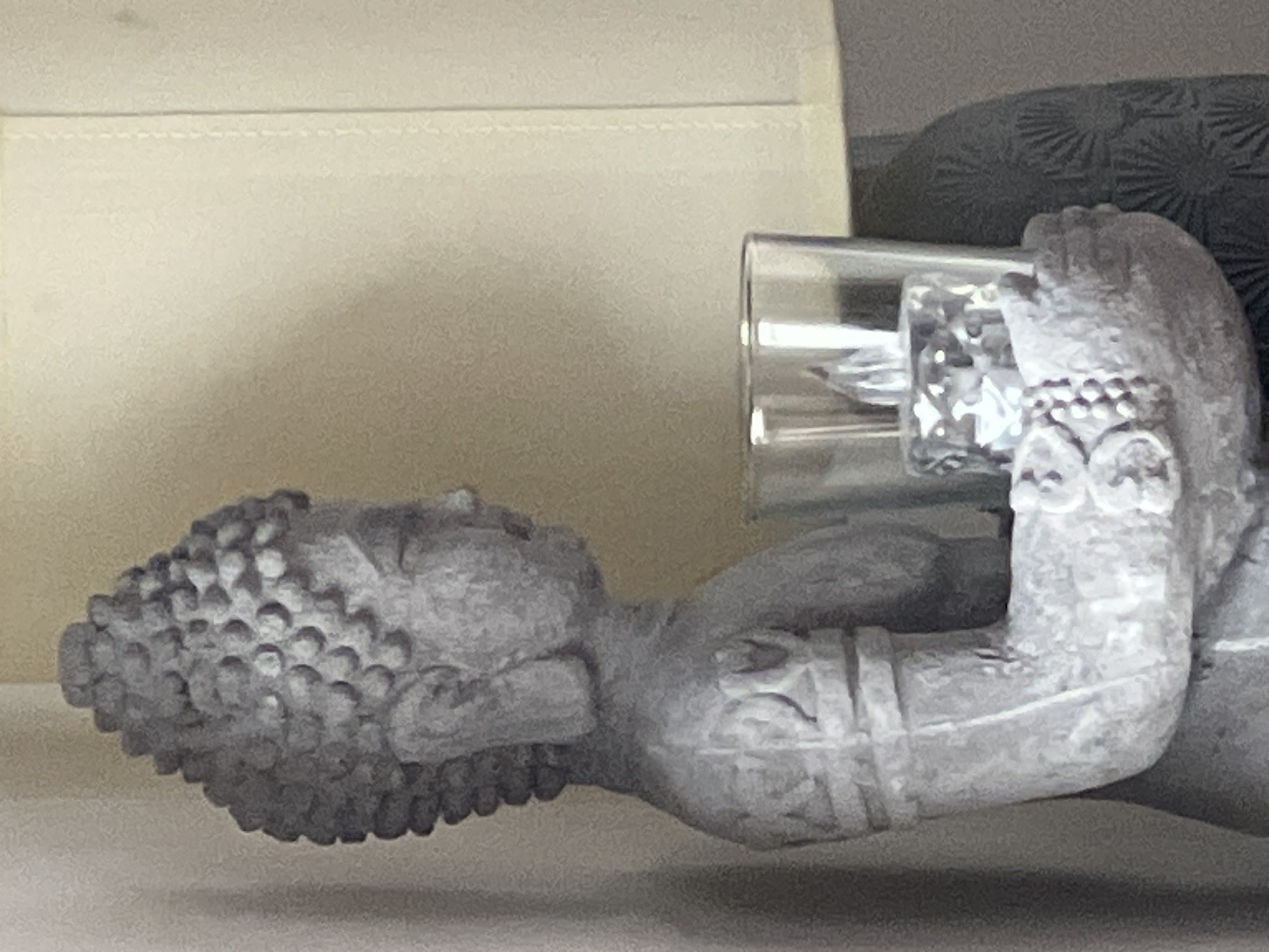 A close-up, side-view shot shows a grey, textured statue of a Buddha figure. The Buddha is holding a clear glass votive holder containing a clear, faceted candle with a visible flame. The candle appears to be electric. The statue's arm and hand are visible, adorned with beaded bracelets and carved bands. The background consists of a plain, cream-colored surface with a visible seam, possibly a lampshade, and a darker grey textured object on the right. The image is an indoor still life, with no discernible location in Bunbeg, Ireland.