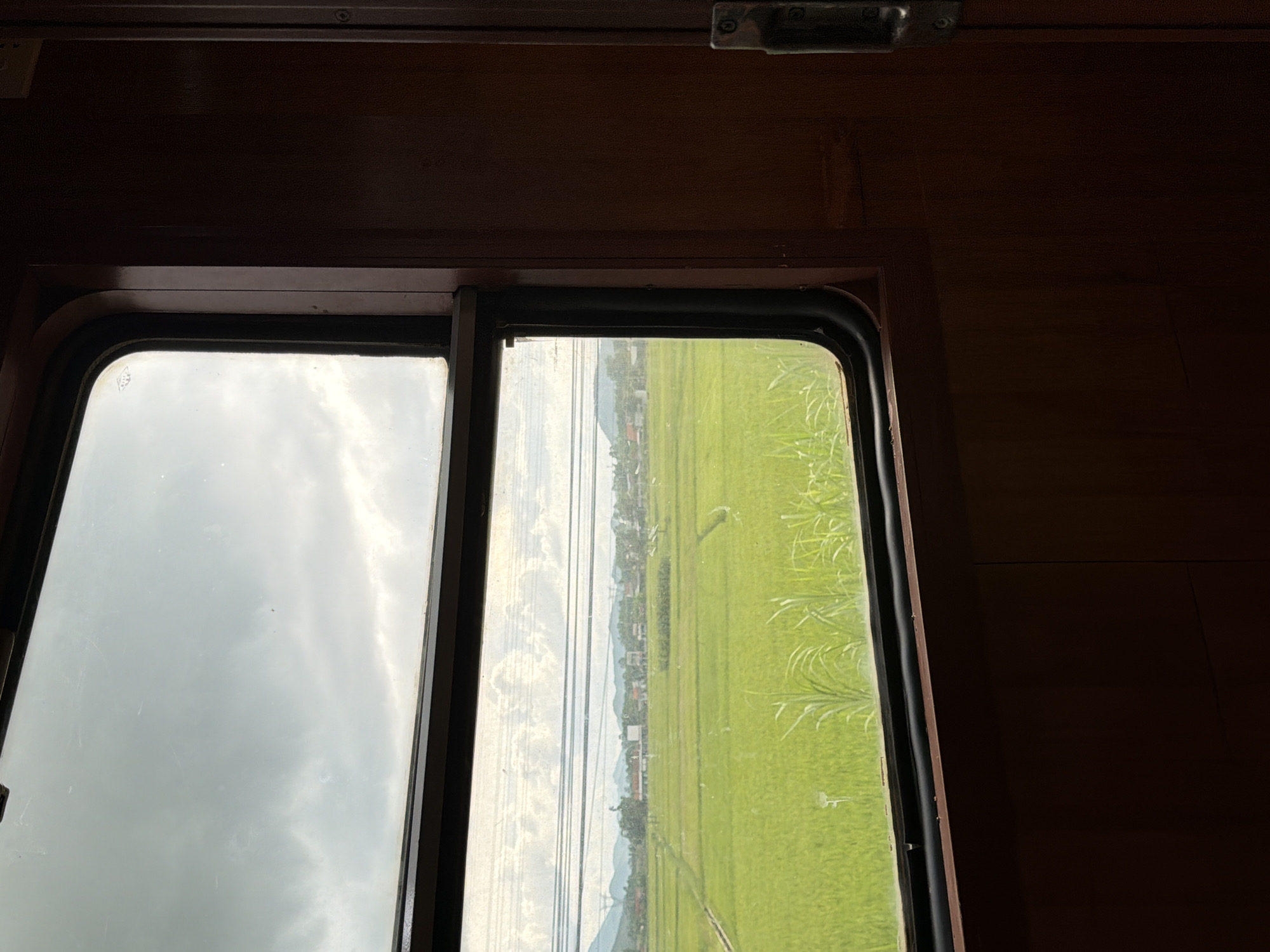 The image shows a view from inside a train carriage, looking out a window at a rural landscape. The window is divided into two panes, with the upper pane showing a cloudy sky and the lower pane revealing a vibrant green rice paddy field stretching towards a distant treeline and a cluster of houses. Power lines are visible in the middle ground, and rolling hills form the backdrop. The interior of the train carriage is characterized by dark wooden paneling, with a visible door handle on the right side of the frame. The scene is devoid of people. Based on the landscape and the typical mode of transportation, this scene could be from a train journey through the outskirts of Da Nang, Vietnam, specifically in an agricultural area surrounding the city.