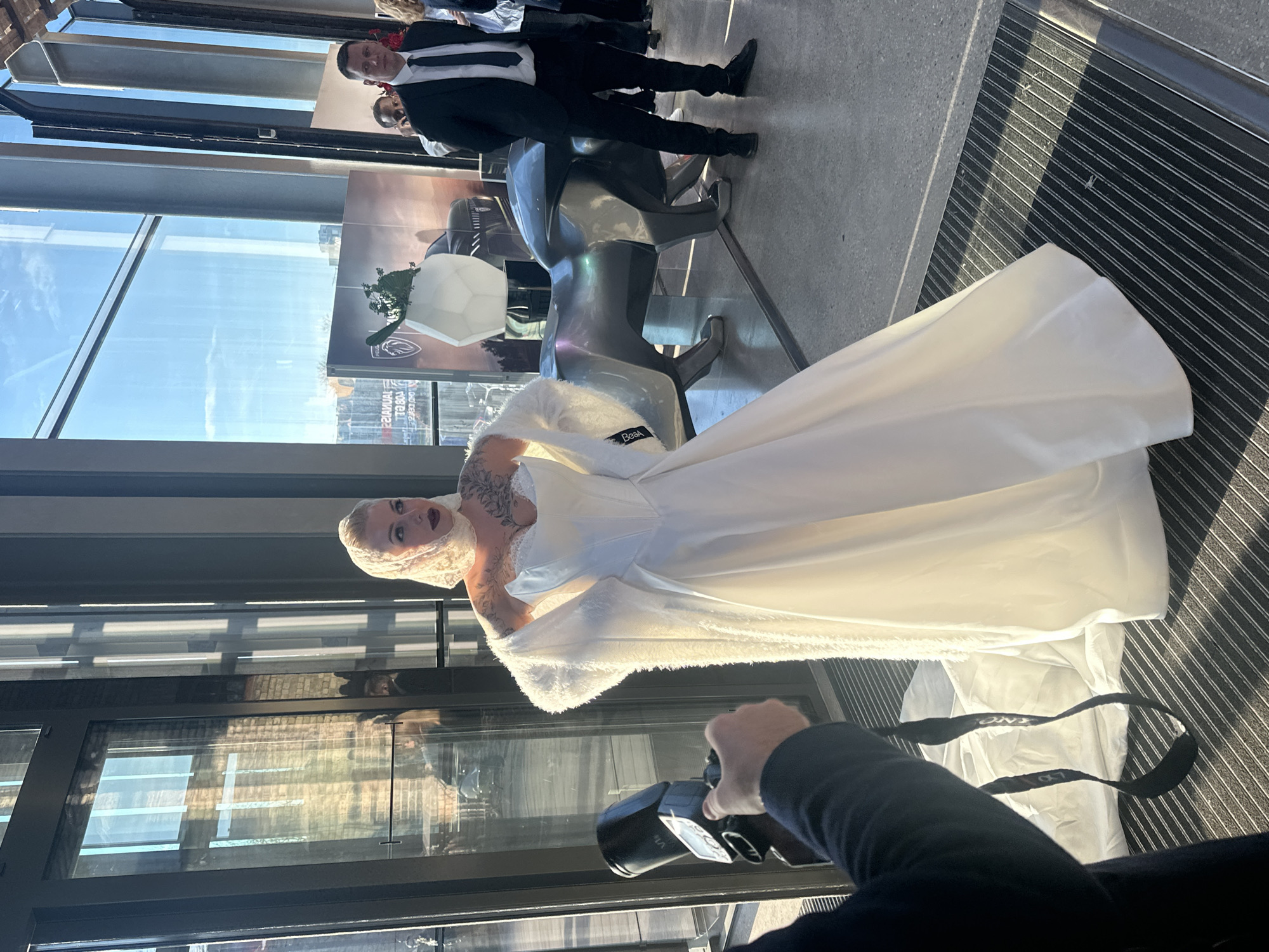 A woman in a white wedding dress poses for a photograph indoors, with a man taking the picture in the foreground. The setting appears to be a modern building with large windows offering a city view, possibly in Riga, Latvia, given the architectural style and landmarks visible in the reflection. The wedding dress has a lace bodice and a long train, and the woman is wearing a lace head covering. The photographer is holding a camera and aiming it at the bride. Other people are visible in the background, including a man in a suit and tie. A futuristic sculpture of a bull stands nearby. The floor is made of a speckled concrete with a dark grill flooring near the edge of the frame. The sky is clear.
