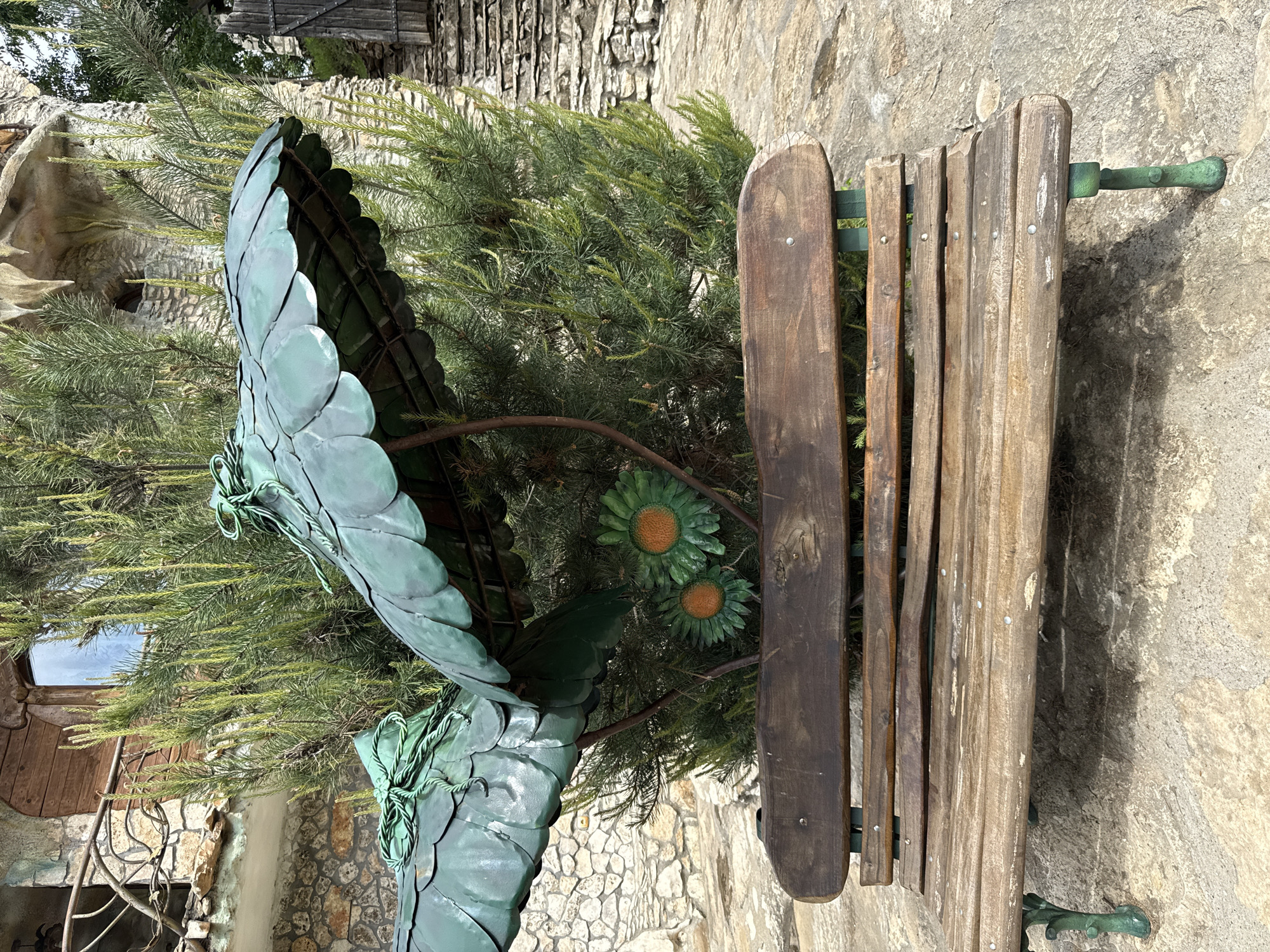 A medium shot shows an outdoor scene featuring a wooden park bench adjacent to a sculpted metal plant. The bench, made of aged wood planks attached to green metal supports, sits on a stone surface that extends into the background. Next to the bench stands a metal sculpture of plant leaves with flowers in green and orange hues. Between the bench and metal sculpture is a small pine tree. The background includes stone walls and parts of buildings, indicative of an urban or park setting in Lukovit, Bulgaria.