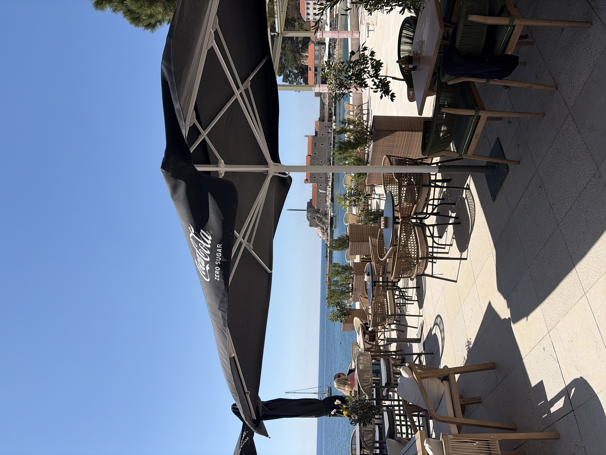 The image shows an outdoor cafe setting on a sunny day in what appears to be a coastal city.
**Location:** Unknown, Unknown
**Environment:** The foreground is dominated by outdoor cafe furniture – tables and chairs, many made of wicker or rattan. Several tables are covered with black umbrellas. The background reveals a body of water and a historic city with stone buildings and red tile roofs. The sky is clear and blue.
**Objects:**
* Cafe tables and chairs
* Coca-Cola branded umbrellas
* City buildings with red tile roofs
* Body of water
**People:**
* There appear to be people seated at the tables near the water, however they are not the main subject of the image.
**Actions/Interactions:** The scene is a static view of a restaurant setting.
