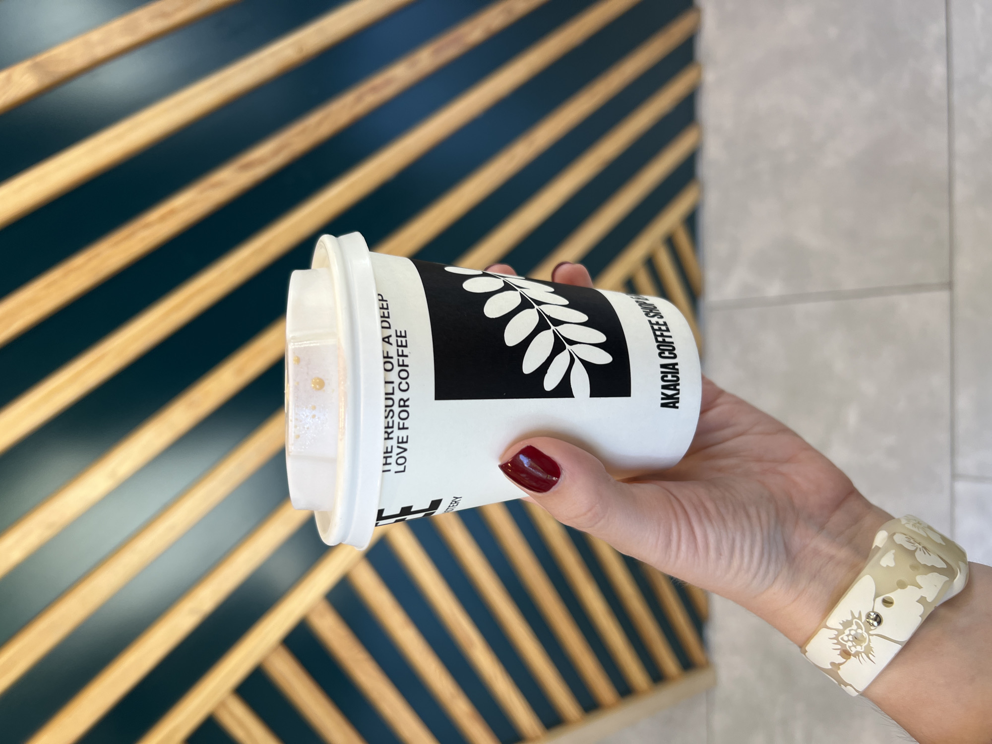 A close-up shot features a person's hand holding a white disposable coffee cup. The hand has manicured nails painted in a deep red color and wears a light-colored wristband or bracelet with an intricate floral pattern. The coffee cup has a distinctive black and white design, including a prominent image of leaves. Text visible on the cup reads, "THE RESULT OF A DEEP LOVE FOR COFFEE" and "AKACIA COFFEE SHOP". There is a small amount of foamy beverage visible at the rim of the cup. The background is composed of a striking pattern of diagonal wooden slats set against a dark green-blue wall, suggesting modern interior decor. To the right, a section of a light grey tiled surface is visible. The activity taking place is simply holding a coffee cup, possibly enjoying a drink in a cafe or building in Budva, Montenegro. The image focuses on the aesthetic of the coffee cup and the decorative background.