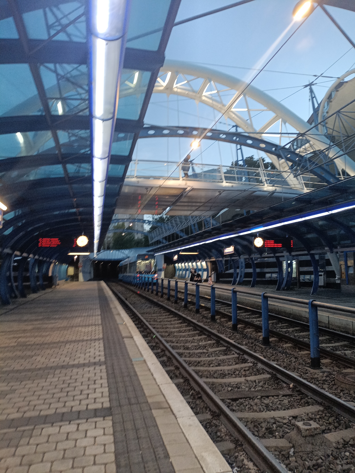 The image shows a modern train station platform in Prague, Czechia. The platform is paved with light-colored bricks and has a textured, darker gray strip along the edge closest to the tracks. On the left side, there is a covered walkway with a blue, curved roof made of glass panels and metal supports, illuminated by long, white fluorescent lights. Red digital displays are visible on the walls of the covered walkway. To the right of the platform are train tracks with ballast. A blue railing runs along the edge of the platform next to the tracks. In the background, there is a large, futuristic-looking arched structure made of white metal and glass, suggesting an open-air section of the station or an adjacent building. The sky is a light blue, indicating it is either early morning or late evening. A train is partially visible in the distance on the right. The overall impression is of a clean, contemporary public transportation hub.