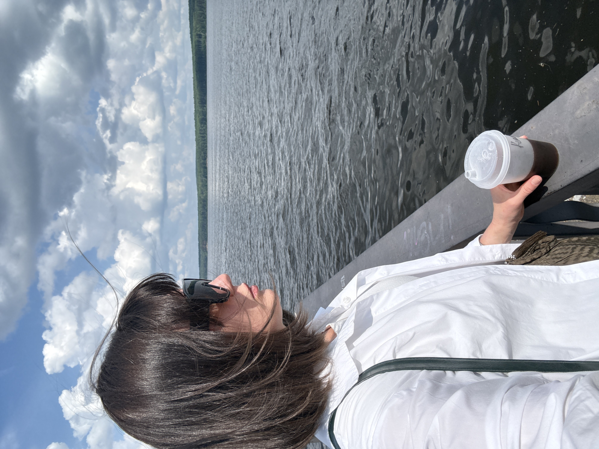 A woman with dark hair and sunglasses stands on a railing overlooking a body of water. She is wearing a white collared shirt and holding a cup with a dark liquid. The water is choppy and reflects the cloudy sky. A treeline is visible in the distance on the far bank. The scene appears to be along the Kama River in Chaikovsky, Russia.