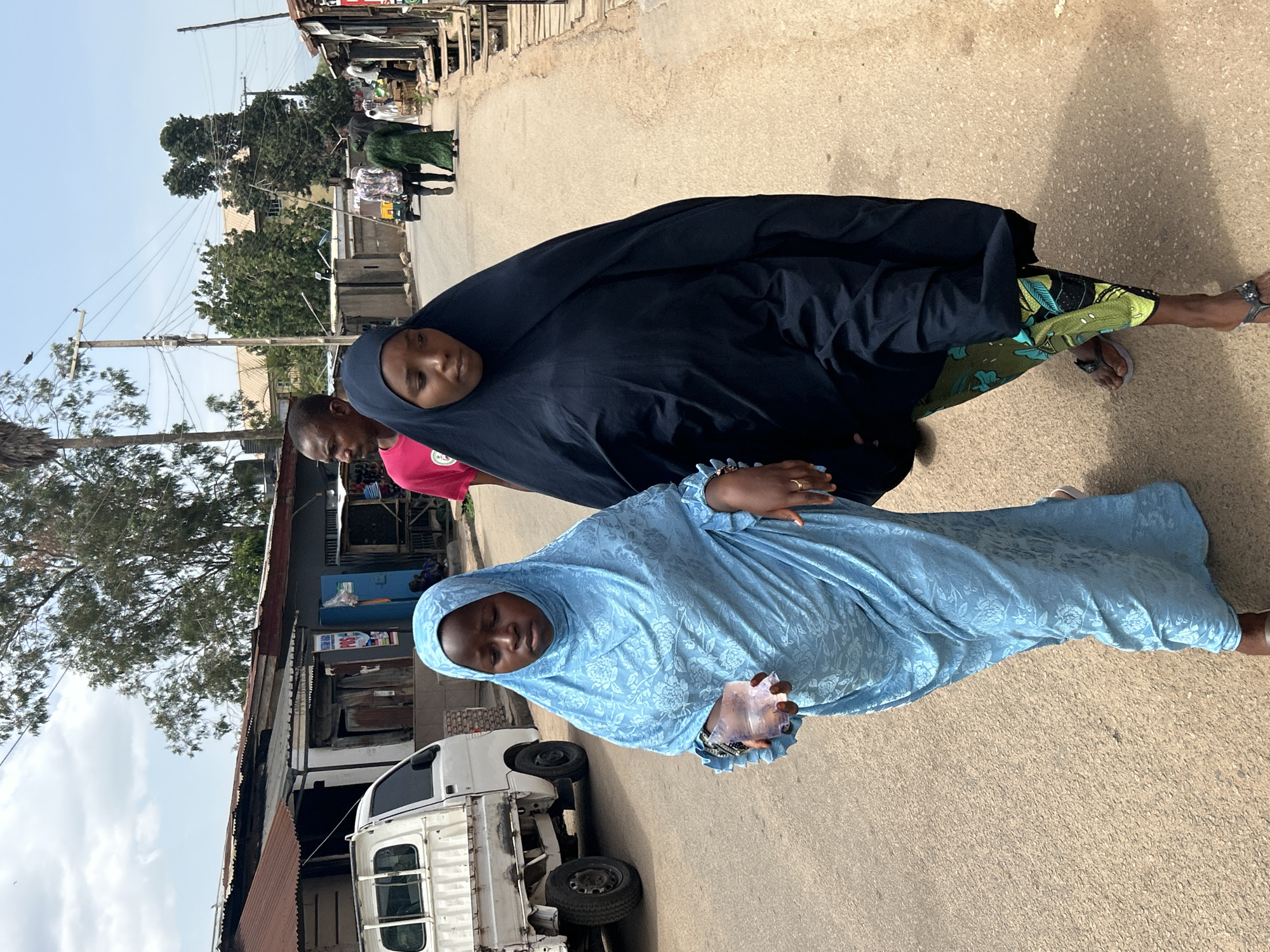 Two women walk along a paved street in Jos, Nigeria. The woman in the foreground wears a blue hijab and dress, holding a plastic bag in her left hand. A white truck is parked nearby. Behind her, another woman wears a black hijab and a patterned green and black skirt. A man in a pink shirt walks behind them. The backdrop includes buildings with corrugated metal roofs, trees, and other people.