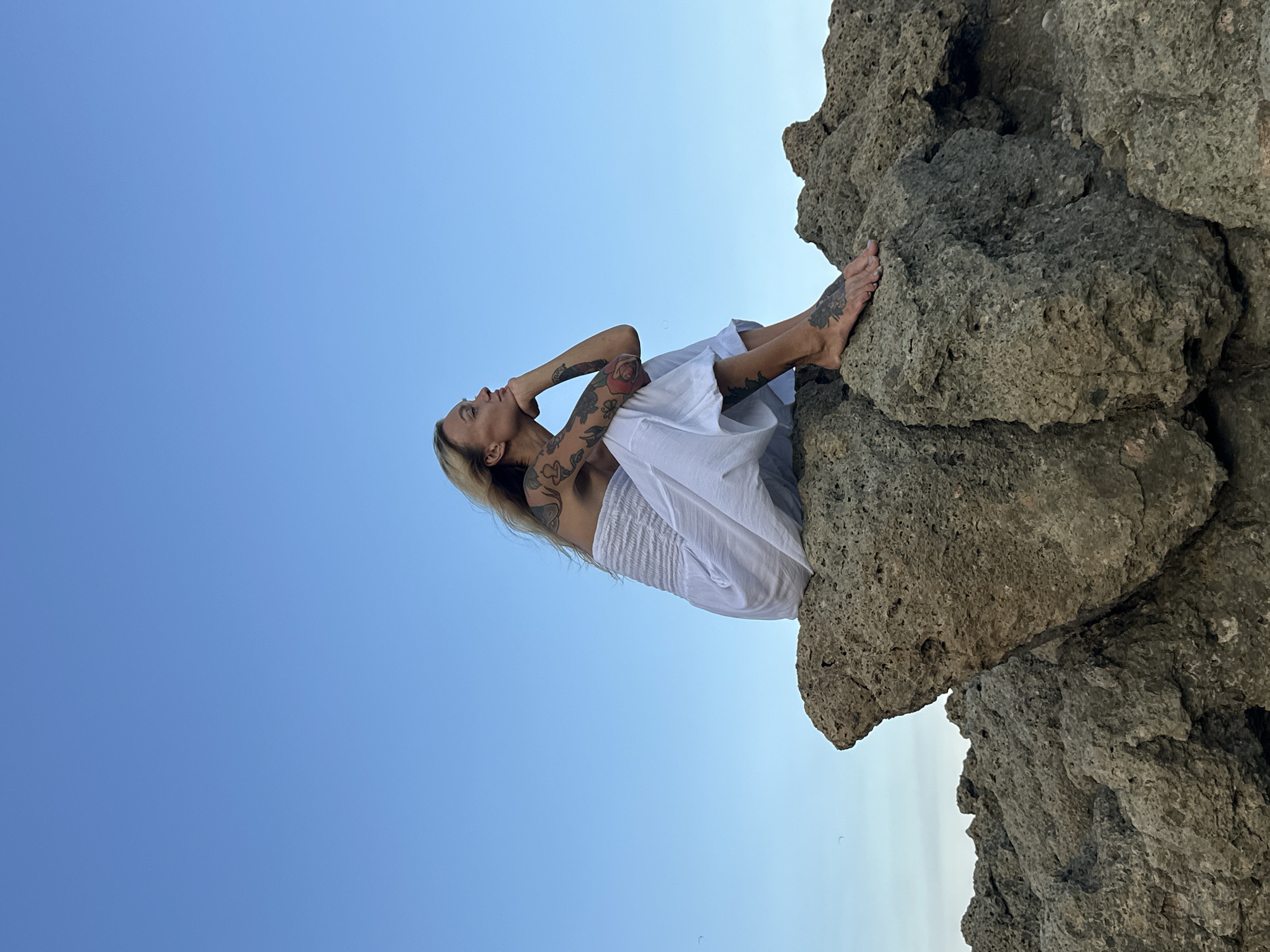 A tattooed woman in a white strapless dress sits on a rocky outcrop, looking out to sea under a clear blue sky. She is resting her chin on her hand and appears to be in deep thought. The rocks are rough and textured, with visible pores and crevices. The woman's body is partially obscured by the rocks, but her arms, legs, and feet are visible, all adorned with intricate tattoos. The ocean is not visible in the frame, but the expansive blue sky suggests an open, possibly coastal, environment. It is not possible to determine the precise location within Mũi Né, Vietnam, based solely on this image.