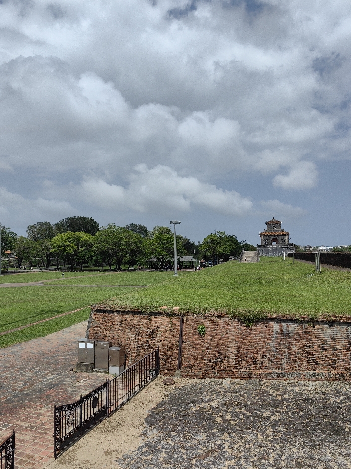 The image depicts an expansive outdoor scene within the Imperial City of Hue, Phú Xuân Ward, Vietnam. The sky occupies the upper two-fifths, featuring dense light gray and white cumulus clouds with patches of clear blue sky.
In the foreground, a dark, rough ground surface covers the lower right. To the left, an ornate black metal fence extends, bordering a reddish-brown brick pathway. Three metallic utility boxes are positioned against a weathered brick wall that runs horizontally across the mid-ground. This brick wall shows signs of discoloration and minor vegetation growth.
Above the brick wall, a large, well-maintained green grassy lawn extends into the background, sloping gently upwards towards the right. A reddish-brown brick path is visible on the far left of the grassy area. A line of dense green trees, interspersed with smaller structures and a tall, multi-headed streetlamp, stretches across the mid-distance. Several small human figures are discernible walking on the grass and near these distant structures.
Prominently situated on an elevated grassy mound on the right side of the image is a traditional Vietnamese multi-tiered pagoda-like structure. It features dark grey walls, a distinctive reddish-orange tiled roof with curved eaves, and a central arched opening. A set of steps leads up to this structure. Faint outlines of distant urban buildings are visible on the far right horizon.