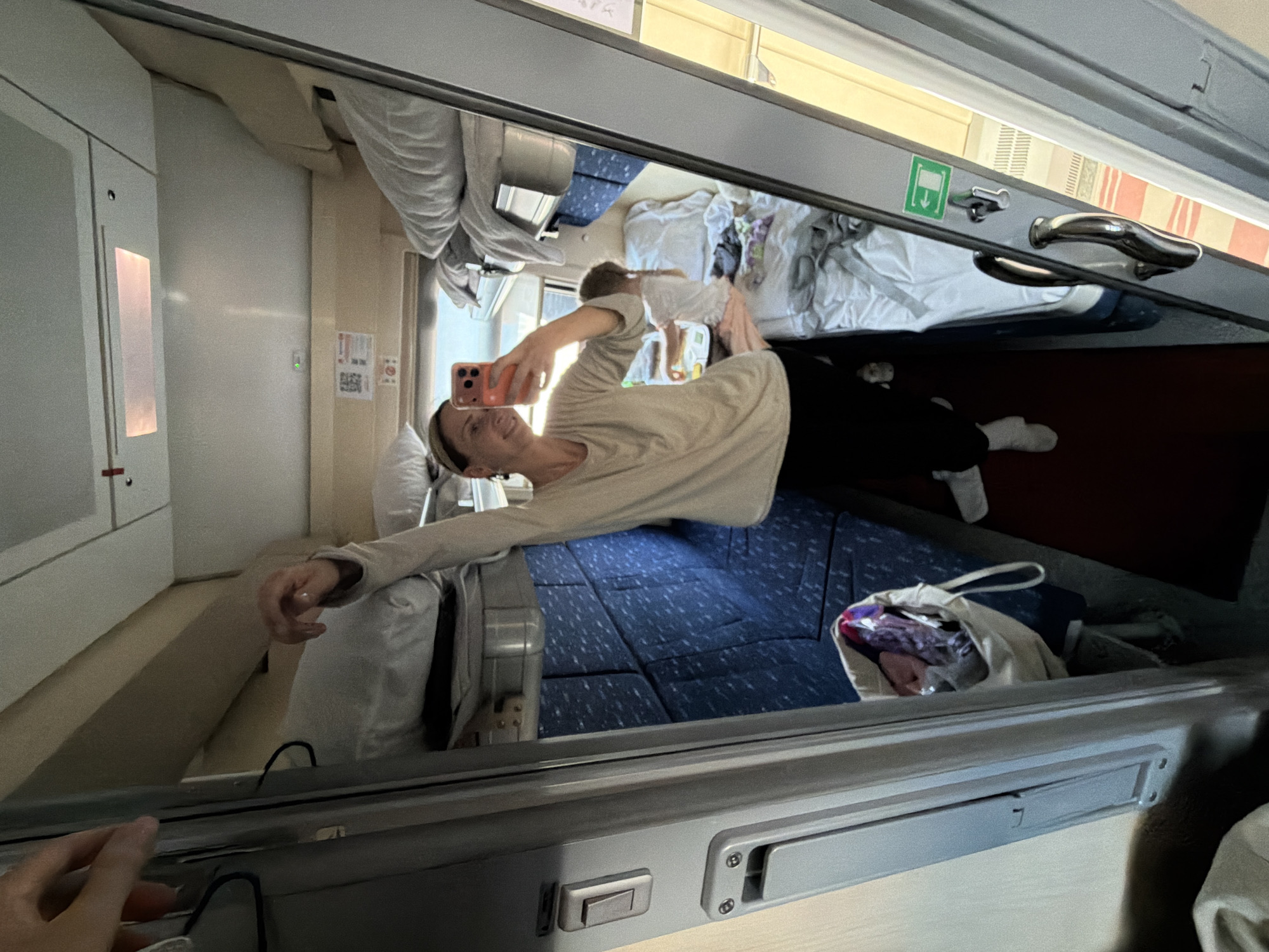 The image captures the reflection of a woman taking a selfie in what appears to be a train compartment. The compartment has bunk beds with blue and white bedding. The woman is holding a smartphone and smiling at the camera. There's another person, possibly a child, in one of the top bunks. A cloth bag is visible near the lower bunk. The location is not provided and is not discernible from the image. The image does not provide any clues that indicate the location is Saratov, Russia.
