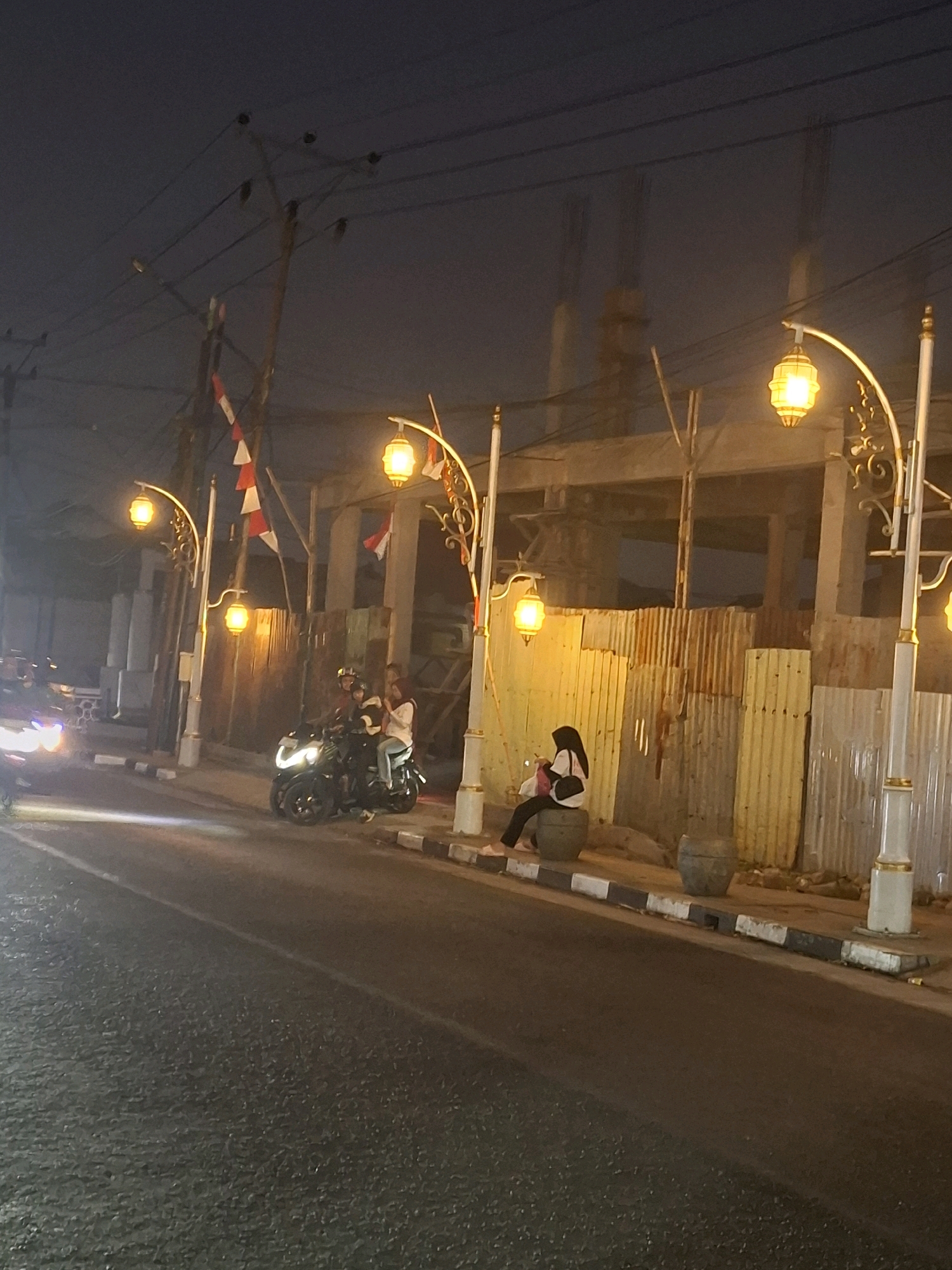 In the city of Sayang, Indonesia, this image captures a street scene at night. The foreground features a dark asphalt road with a white line marking its edge. To the right, a sidewalk with a black and white striped curb is visible, leading to a textured wall constructed from corrugated metal sheets, predominantly in yellow and rust tones.
Several ornate streetlights with golden lanterns illuminate the scene. Two individuals are seated on a round, stone-like structure on the sidewalk. One person is wearing a black hijab and a light-colored top, holding a bag. The other figure is not clearly discernible.
Behind them, a three-wheeled motorcycle with two passengers is parked. The driver and a passenger in the back are wearing helmets. The passenger in the middle is wearing a light-colored top.
In the background, a building under construction is partially visible, with exposed concrete structures and scaffolding. Indonesian flags, red and white, are strung up along a utility pole on the left side of the image. Another motorcycle is seen in the far left, its headlights illuminated, suggesting it is either approaching or moving away from the camera. The sky is dark and hazy, typical of a night in a built-up area.