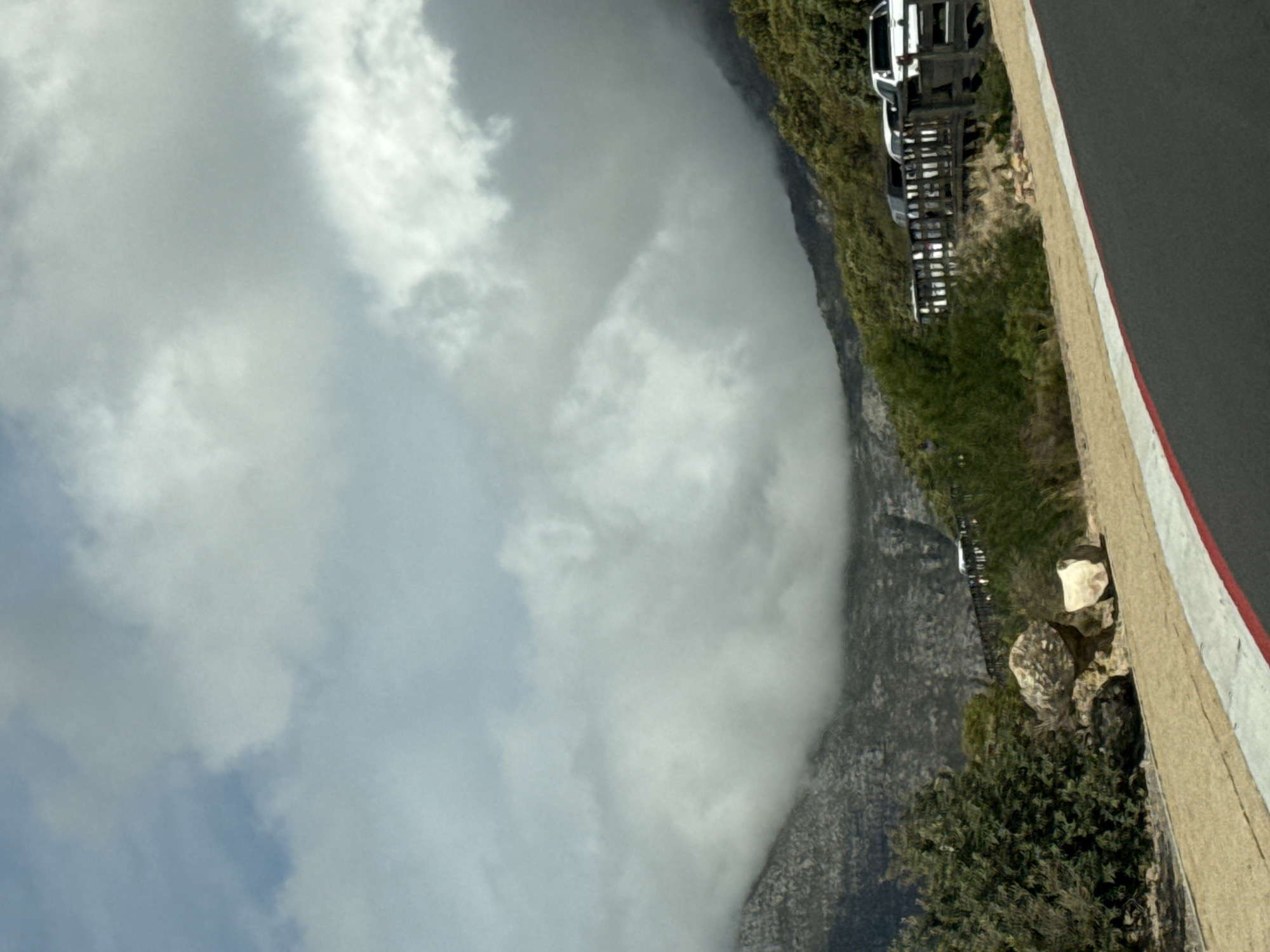 The image shows a view of Table Mountain in Cape Town, South Africa. The mountain is partially obscured by heavy clouds. A road runs along the side of the mountain, with several vehicles parked on it. There are also rocks and vegetation visible along the roadside.