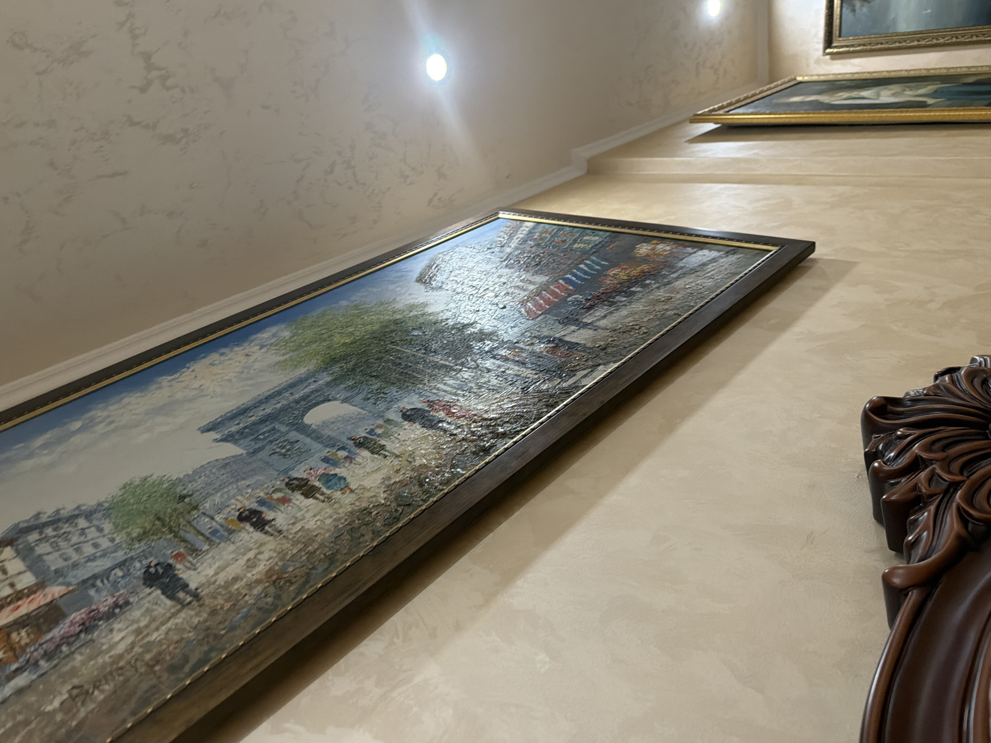 A large oil painting of a Parisian street scene hangs on a textured, beige wall. The painting, framed in dark wood with a gold inner trim, depicts people walking past buildings and an archway under a cloudy sky. The artwork is angled, showing its side and a portion of the wall, which has subtle, darker textured patterns. To the right, a second framed painting with a gold frame is visible, and further to the right, a third painting with a similar gold frame is partially visible. The ceiling, also beige and textured, features a white molding and a bright recessed light fixture. In the lower left corner, the ornate, dark wood carving of a piece of furniture is visible. There is no identifiable information in the image to suggest a location in Vesyoloye, Russia.