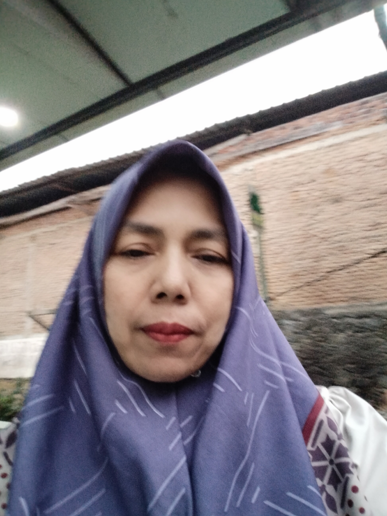 An outdoor, eye-level, medium-shot selfie shows a woman wearing a purple hijab with a white geometric pattern. Her head is slightly tilted, and she is looking at the camera with her right eye slightly squinted. Her lips are painted red, and her eyebrows are dark and arched. The background consists of a brick wall with a corrugated metal roof above it. A thin, green vertical object is visible on the brick wall behind her. The image appears to have been taken in a residential or semi-urban area.