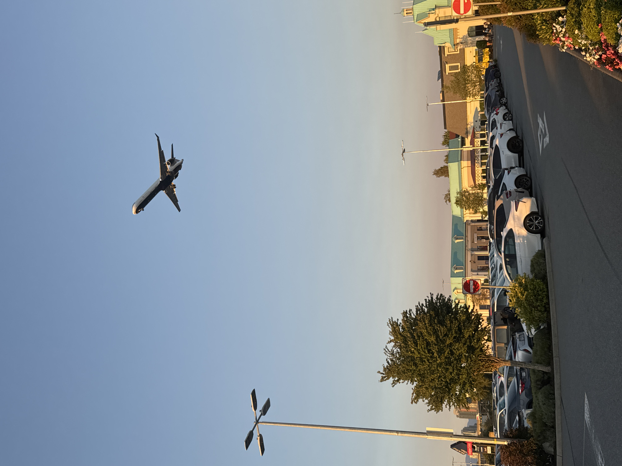An airplane is captured in mid-flight against a clear blue sky. Below, a parking lot is filled with various vehicles. To the left, a tall lamppost with four lights stands prominently. In the middle ground, a lush green tree is visible, surrounded by bushes. Behind the tree and cars, a row of buildings with light blue roofs and yellow facades stretches across the landscape. To the right, a "no entry" sign is posted on a pole. The scene appears to be a sunny day, likely in the late afternoon given the lighting. The location is the Vancouver International Airport (YVR) in Richmond, Canada, which is known for its proximity to the airport and its outlet mall.