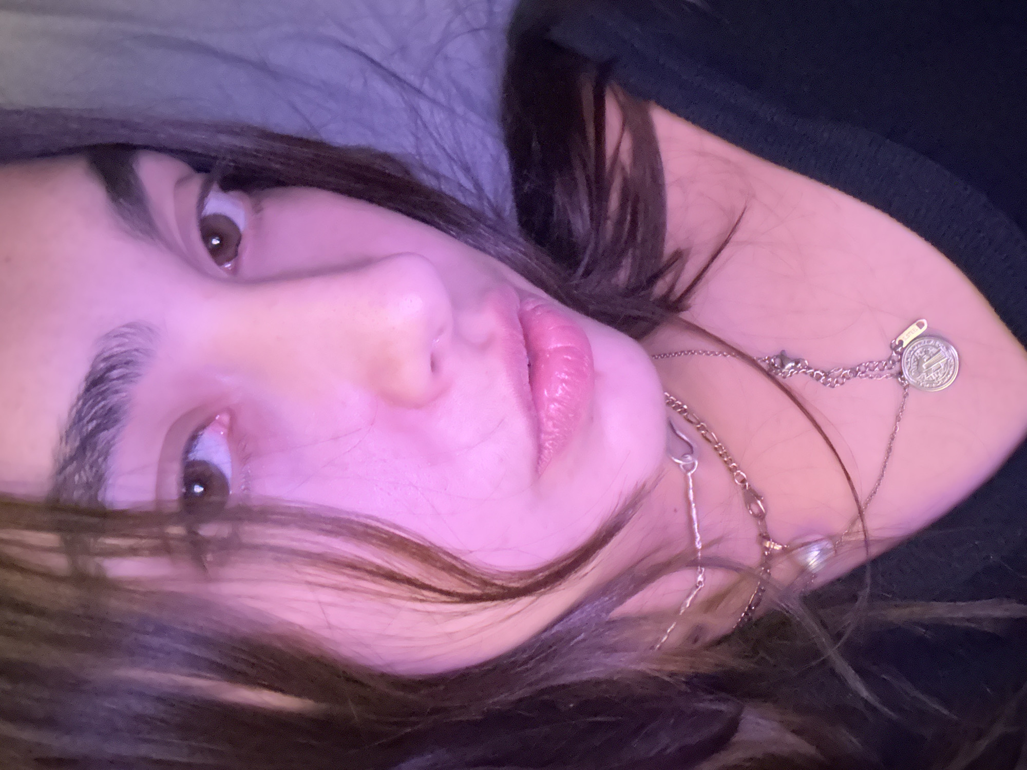 A close-up, tilted portrait captures a young adult female looking directly at the viewer. Her dark brown hair cascades around her face and shoulders, partially obscuring her form. She wears a dark-colored top and two layered silver-toned necklaces. One necklace features a small, round pendant, possibly resembling a coin with a classical design, while another includes a rectangular tag clearly marked "S.STEEL."
The lighting is a striking feature, casting a pervasive pink and purple glow over her skin and the surrounding area, suggesting artificial, perhaps neon or LED, illumination, likely at night or indoors. Her lips are slightly parted, and her expression is neutral and direct. The background is out of focus, appearing as a soft, dark fabric, possibly a blanket or pillow, contributing to an intimate setting. The scene provides no specific architectural or environmental cues, but the context indicates the location is Mexico City, Mexico.