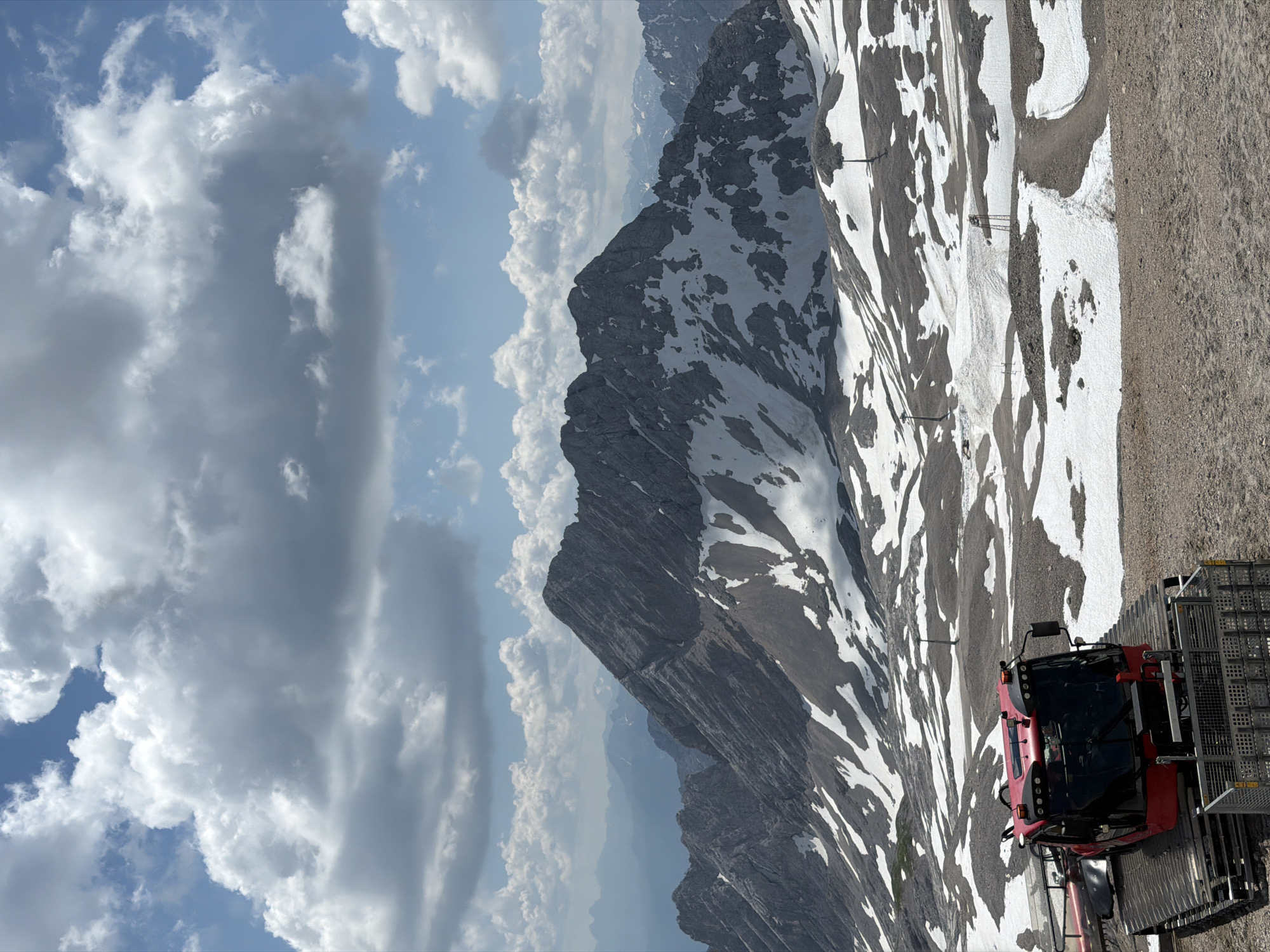 The image depicts a snow-covered mountain range under a partly cloudy sky, likely taken in the Garmisch-Partenkirchen region of Germany, known for its alpine scenery. In the foreground, a red snow groomer vehicle is parked on a gravelly surface. The vehicle has a large cabin with tinted windows and tracks for traversing snowy terrain. Behind the snow groomer, the landscape opens up to reveal a vast mountainous expanse. Patches of snow are scattered across the slopes, interspersed with rocky outcrops and dirt paths. Several ski lift towers are visible in the distance, suggesting this area is used for winter sports. The mountains in the background are rugged and imposing, with some peaks still retaining significant snow cover. The sky above is a mix of bright blue patches and large, fluffy white clouds, indicating fair weather with some cloud cover. The overall scene conveys a sense of a high-altitude, mountainous environment, possibly during the late spring or early summer when snow still lingers on the peaks.
