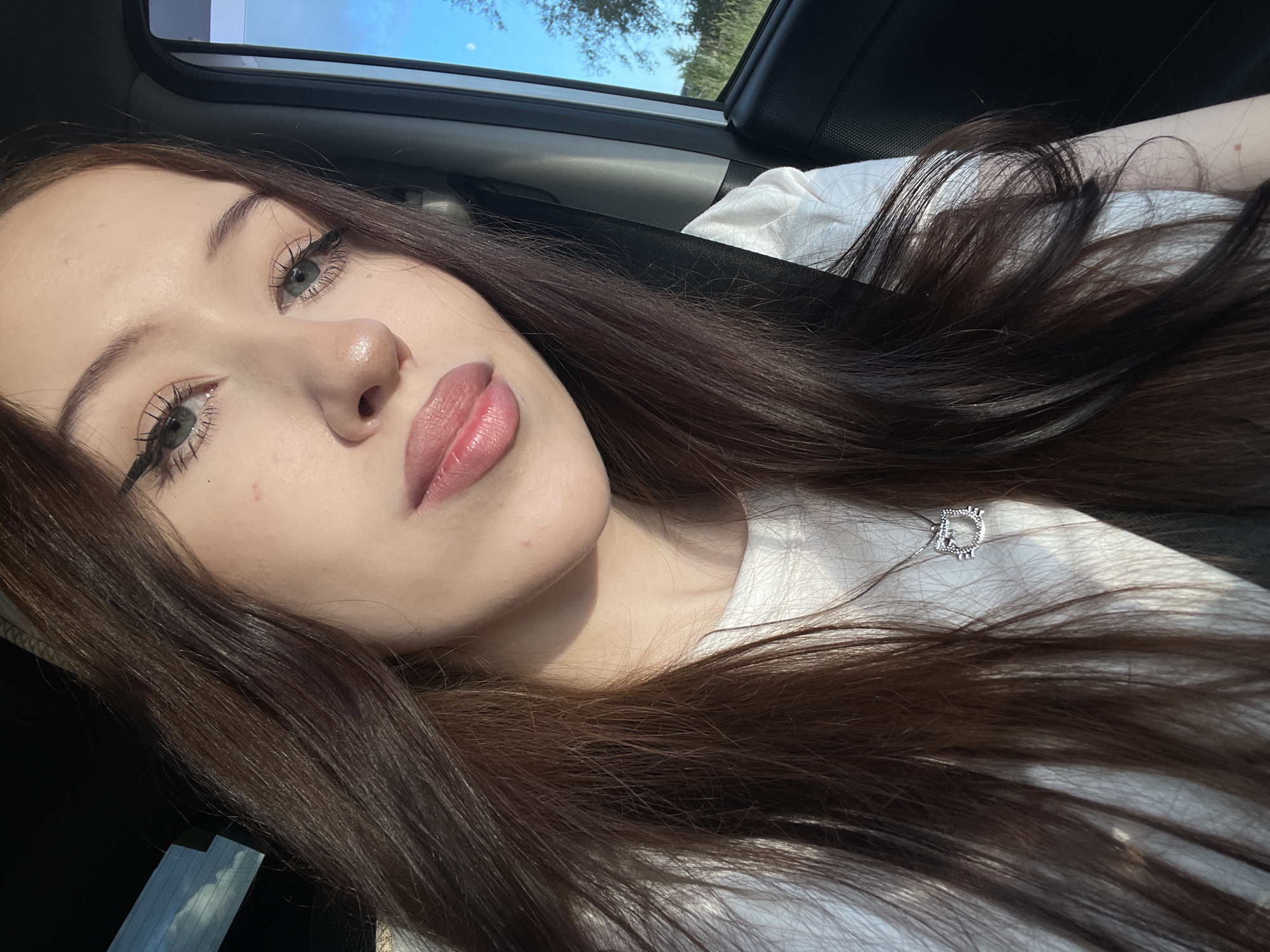 This is a close-up, selfie-style image of a young woman inside a vehicle, likely a car, as suggested by the seatbelt and interior. The woman has long, dark brown hair that is flowing loosely. She is wearing a white t-shirt and a silver pendant necklace with a heart shape. Her makeup includes winged eyeliner and lip gloss. The lighting suggests it is daytime, with sunlight casting shadows and highlights on her face and hair. The background shows a glimpse of the sky and trees through the car window, indicating an outdoor setting. The overall mood is relaxed and casual. There is no visible text in the image.