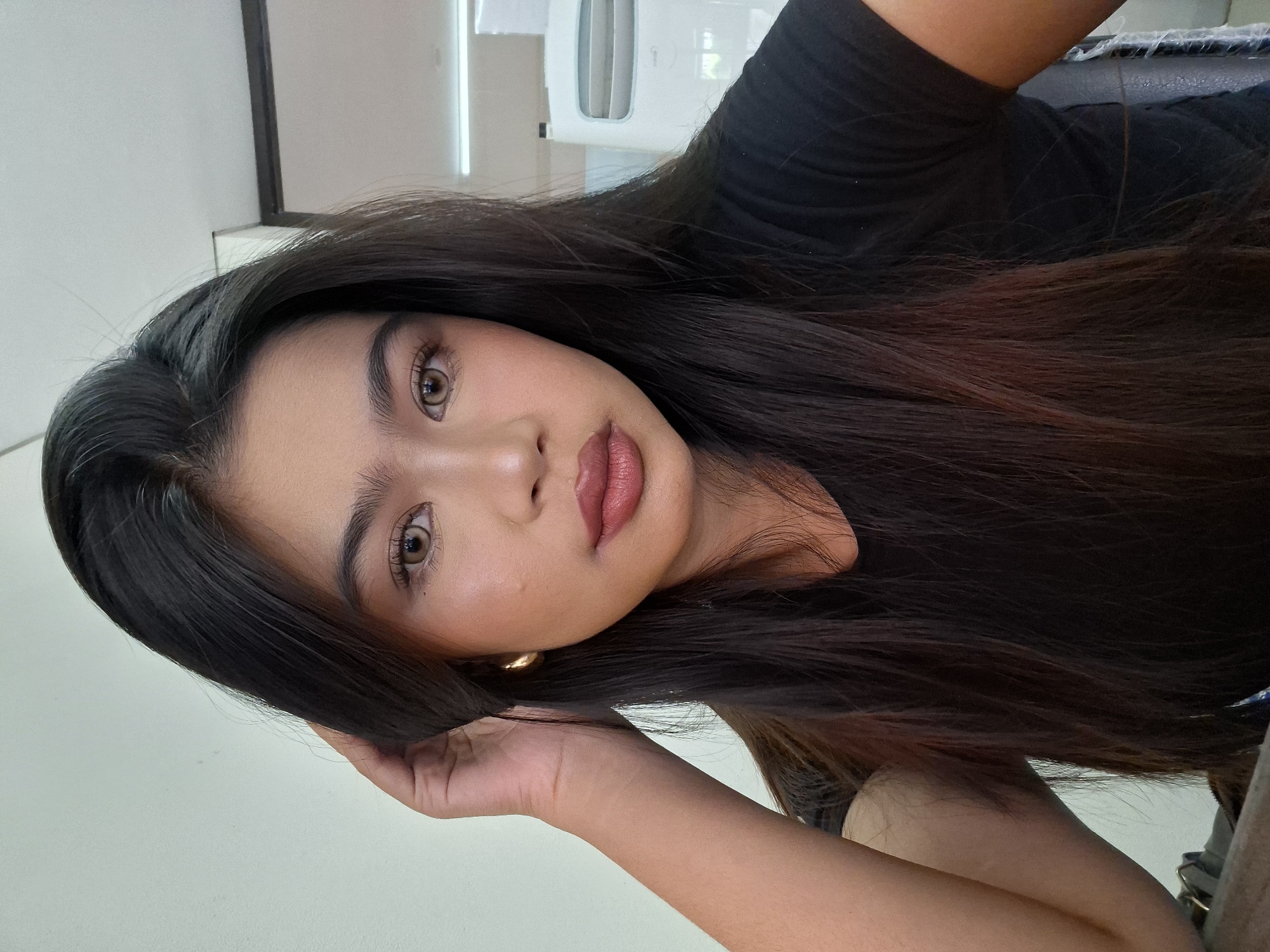 In Kuta, Indonesia, a young woman with long, dark hair is captured in a close-up, head-and-shoulders selfie. She is wearing a black t-shirt and a golden hoop earring. Her left hand is raised, gently touching her hair near her temple. Her makeup includes subtle eyeshadow, defined eyebrows, and a pink lipstick. The background is a plain wall, suggesting an indoor setting.