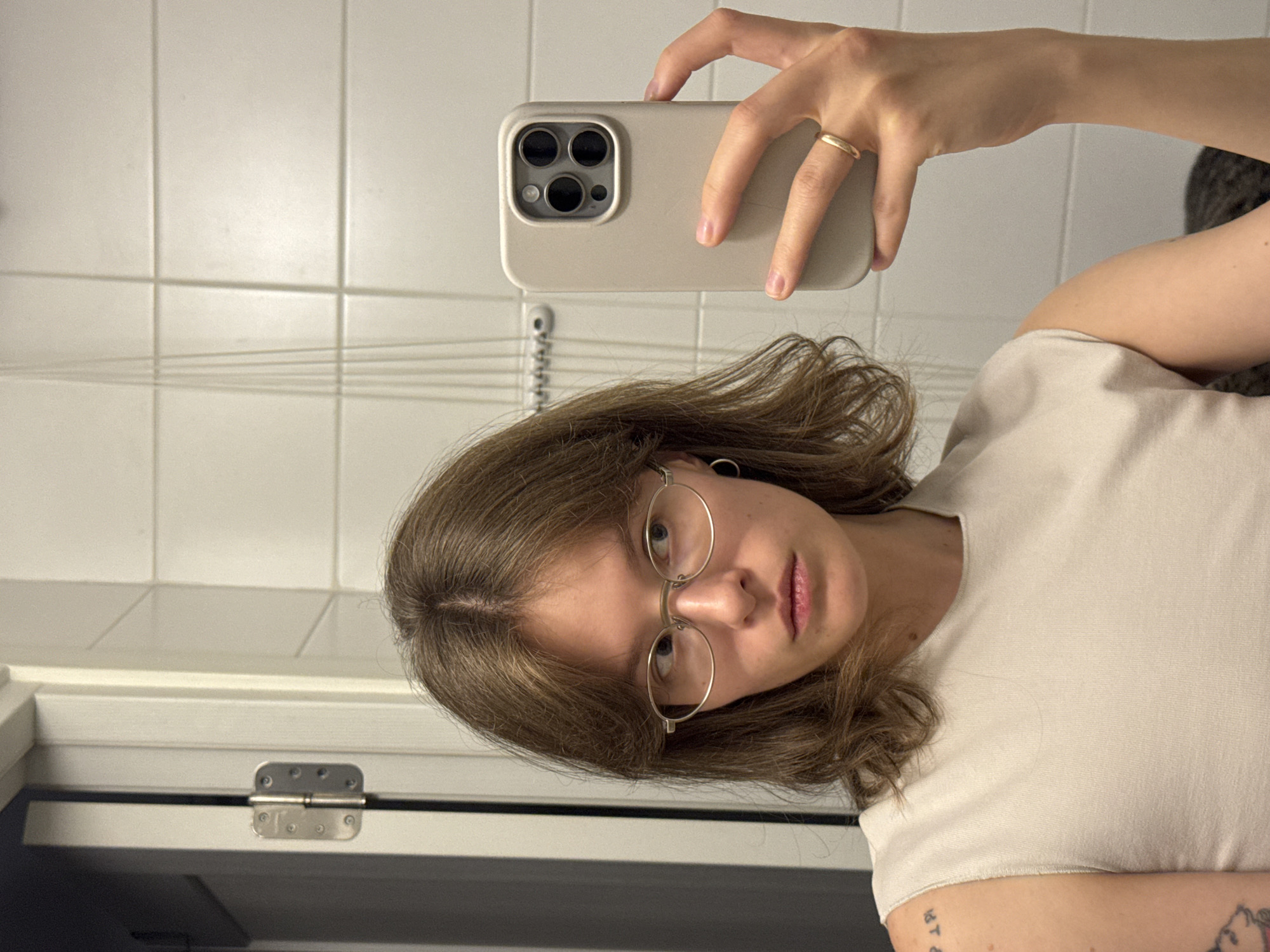 This is a waist-up, self-portrait photograph of a young woman with shoulder-length brown hair and fair skin, taken in a bathroom. She is wearing round, thin-rimmed glasses and a light-colored, sleeveless top. She is looking to her right, away from the camera. Her left hand is visible on her shoulder, showing a tattoo. Her right hand is holding a beige smartphone, angled to capture the reflection in a mirror. A gold ring is visible on her right ring finger. The background consists of white tiled walls and a partially visible white door with a silver hinge on the left side of the frame. There are thin, vertical lines in the background, possibly strings or wires, and a rack with several hooks is mounted on the wall. There is no indication that this image was taken in Moscow, Russia.