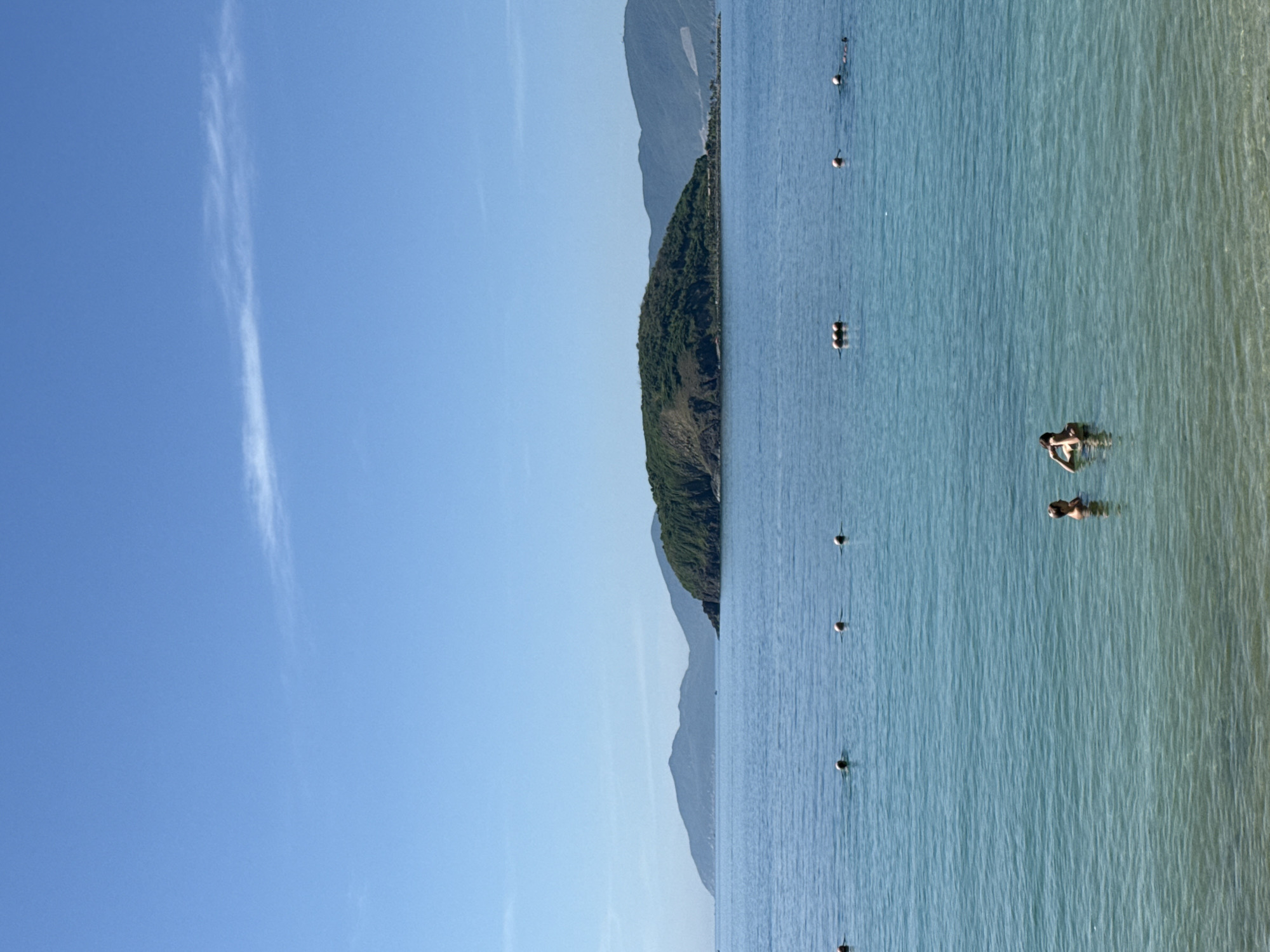 The image depicts a serene beach scene in Nha Trang Ward, Vietnam. In the foreground, the clear, turquoise water laps gently at the shore. Several buoys are spaced out across the water, marking a boundary or swimming area. Two individuals are visible wading in the water, appearing to be enjoying the ocean. In the middle ground, a lush, green island rises from the sea, its slopes covered in vegetation. The island is positioned to the right of the frame. Further in the background, a series of hazy, blue mountains form a distant horizon line under a clear, bright blue sky with a single wispy white cloud. The overall atmosphere is tranquil and inviting, suggesting a pleasant day at the beach.