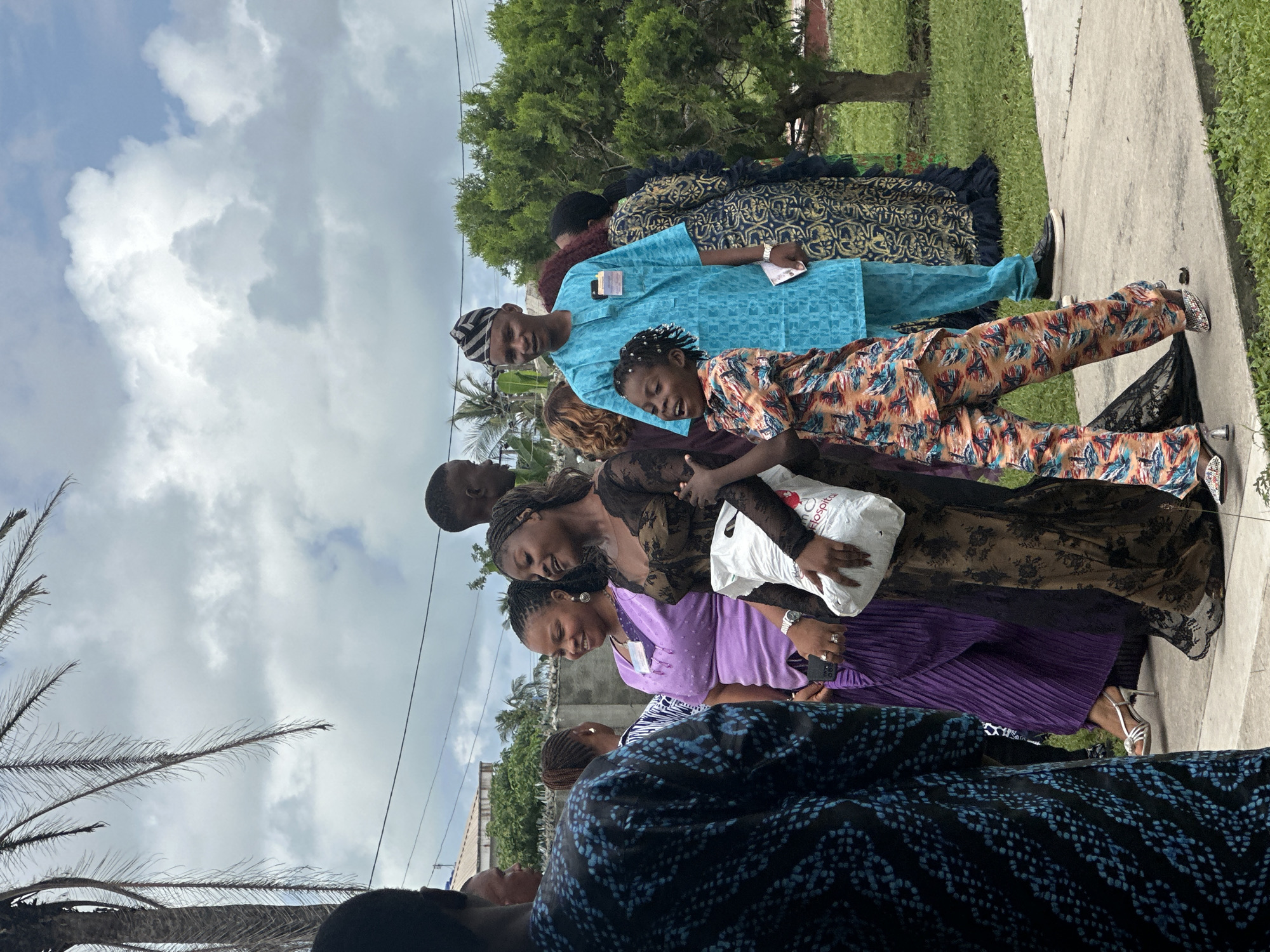 In Itoga, Nigeria, a group of people are gathered outdoors on a cloudy day, possibly for a celebration or event. A woman in a purple dress and a woman in a dark lace dress are talking and smiling. The woman in the lace dress is holding a white bag with a logo that reads "Elegance Hospitals". To their right, a man in a bright blue traditional outfit, wearing a patterned headwrap, stands next to a young child in a colorful patterned jumpsuit. The child is laughing and looking up. In the foreground, a person in a dark blue and black patterned garment is partially visible. Behind the group, a palm tree and lush green vegetation are present. A fence and part of a building are visible in the background, suggesting a residential or community setting.